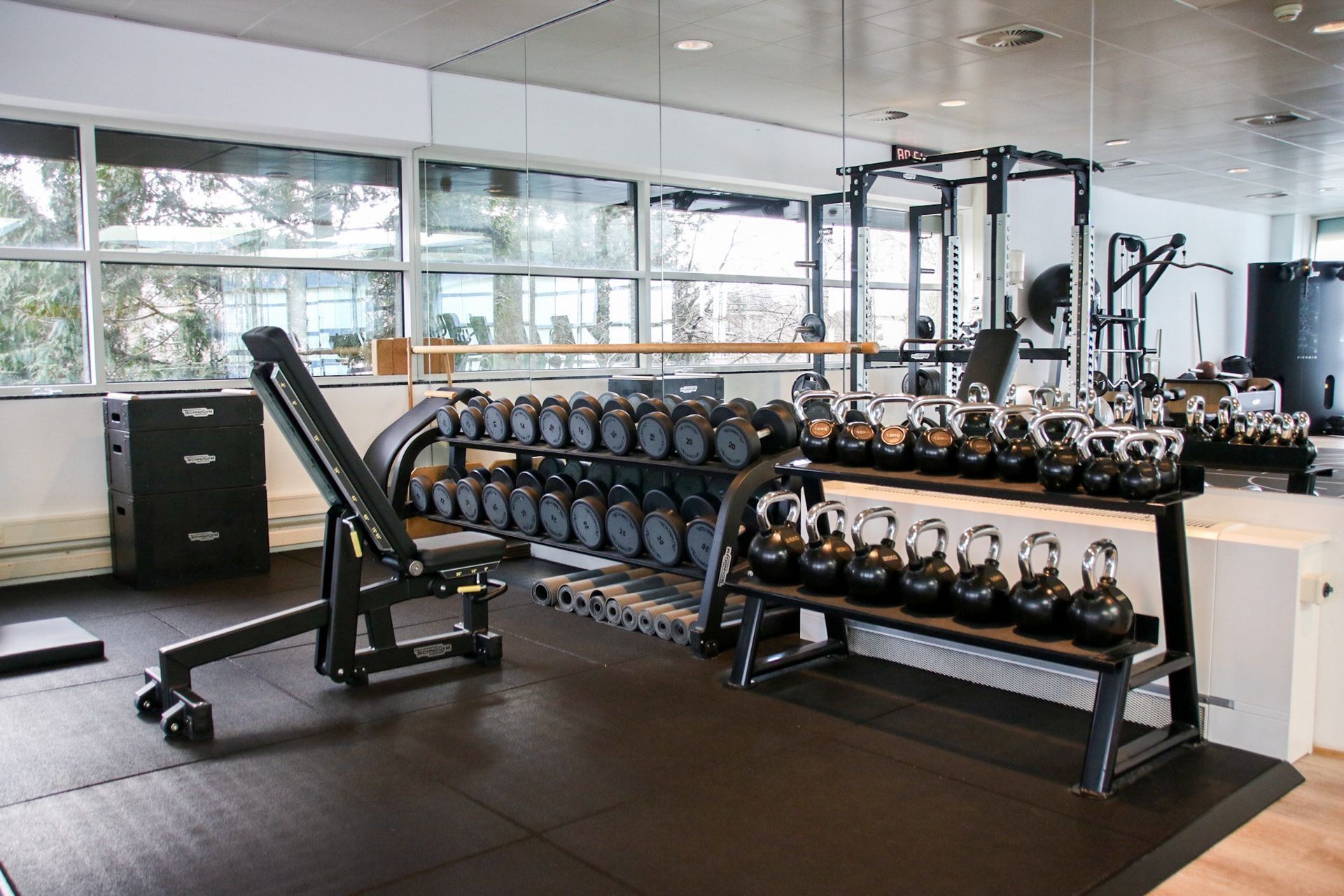 Gym with weights, bench, and training equipment. Dark flooring, light walls, and windows with outdoor view.