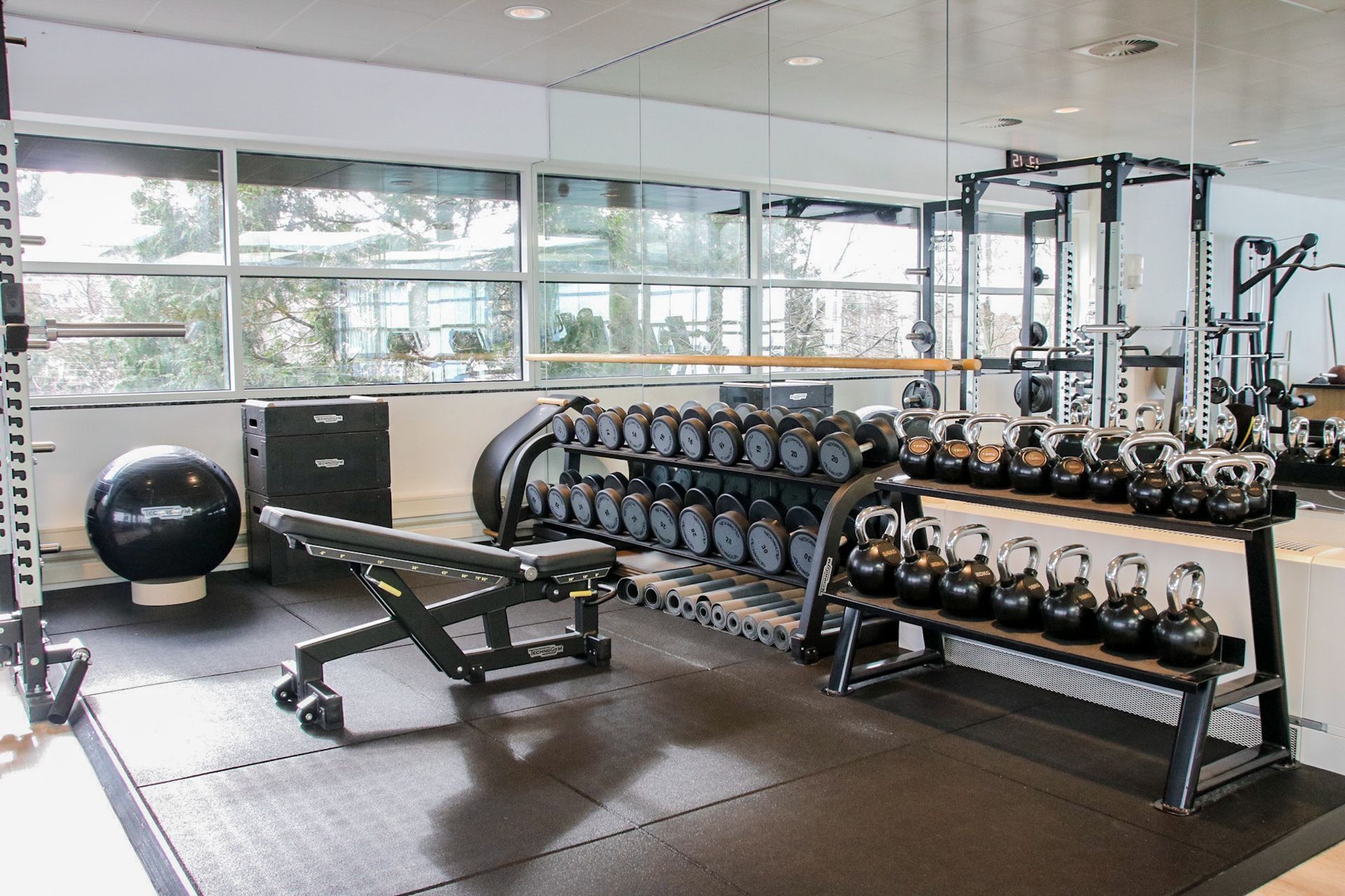 Gym with weights, bench, and equipment against a mirrored wall and large windows.