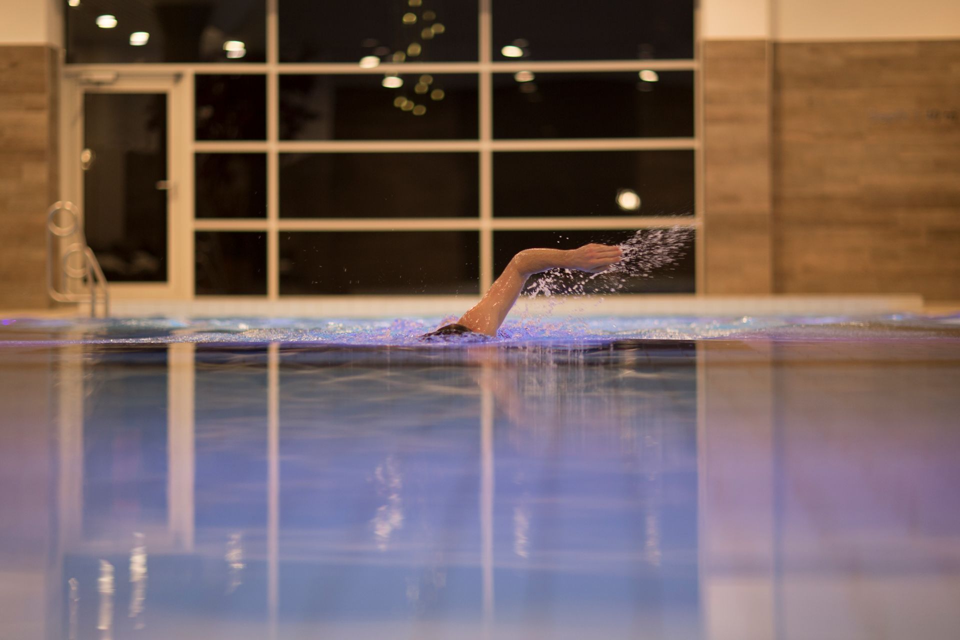 A swimmer's leg emerges from pool water, kicking up a spray. Indoor pool with a large window.
