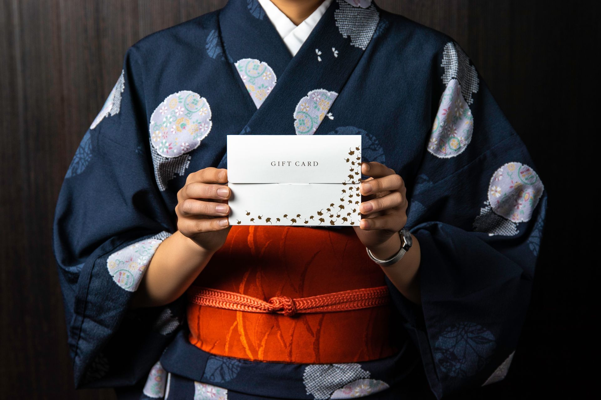 Person in yukata holding a gift card, orange sash, dark background.
