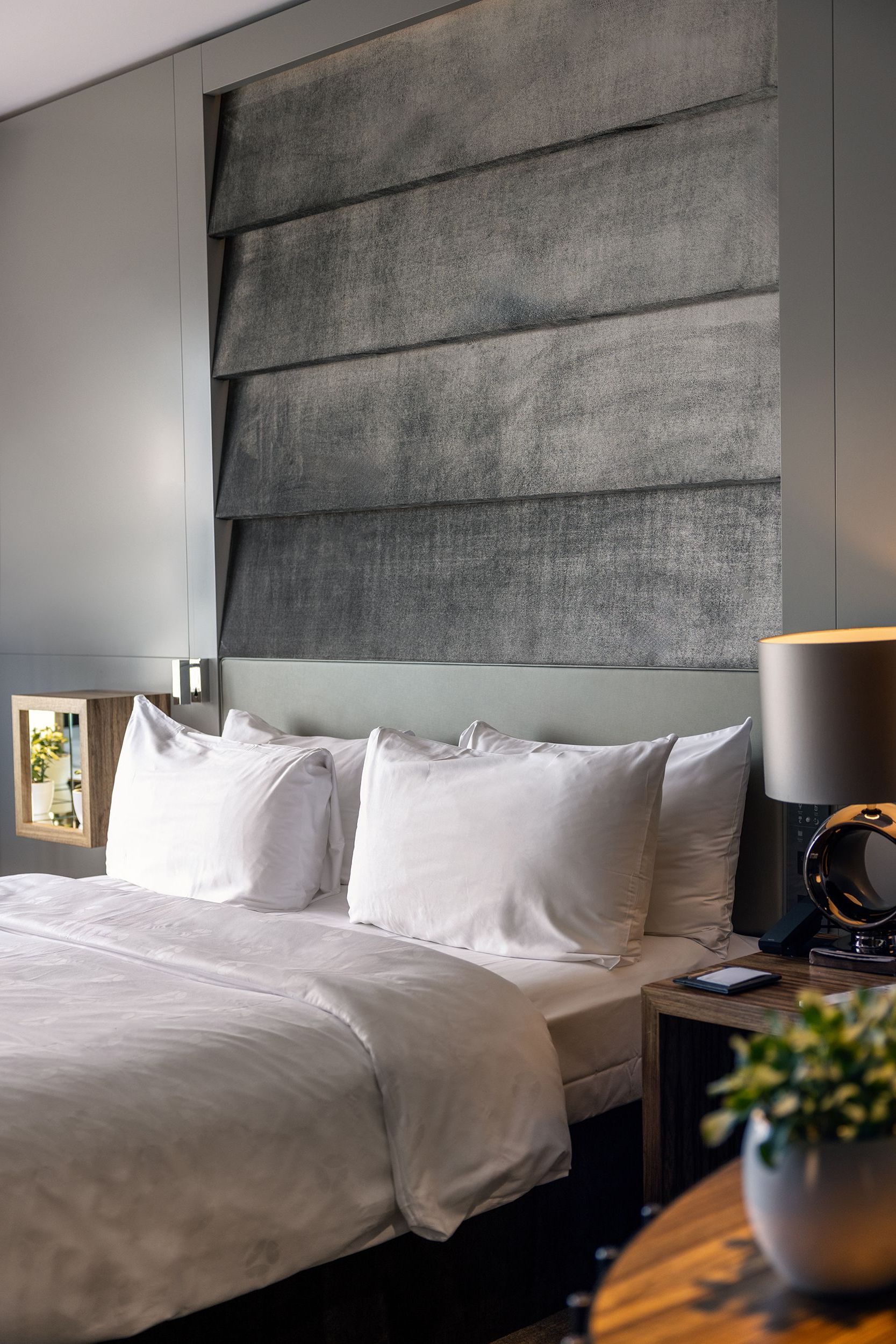 Bedroom with white bedding, gray headboard, bedside table with lamp and plant.