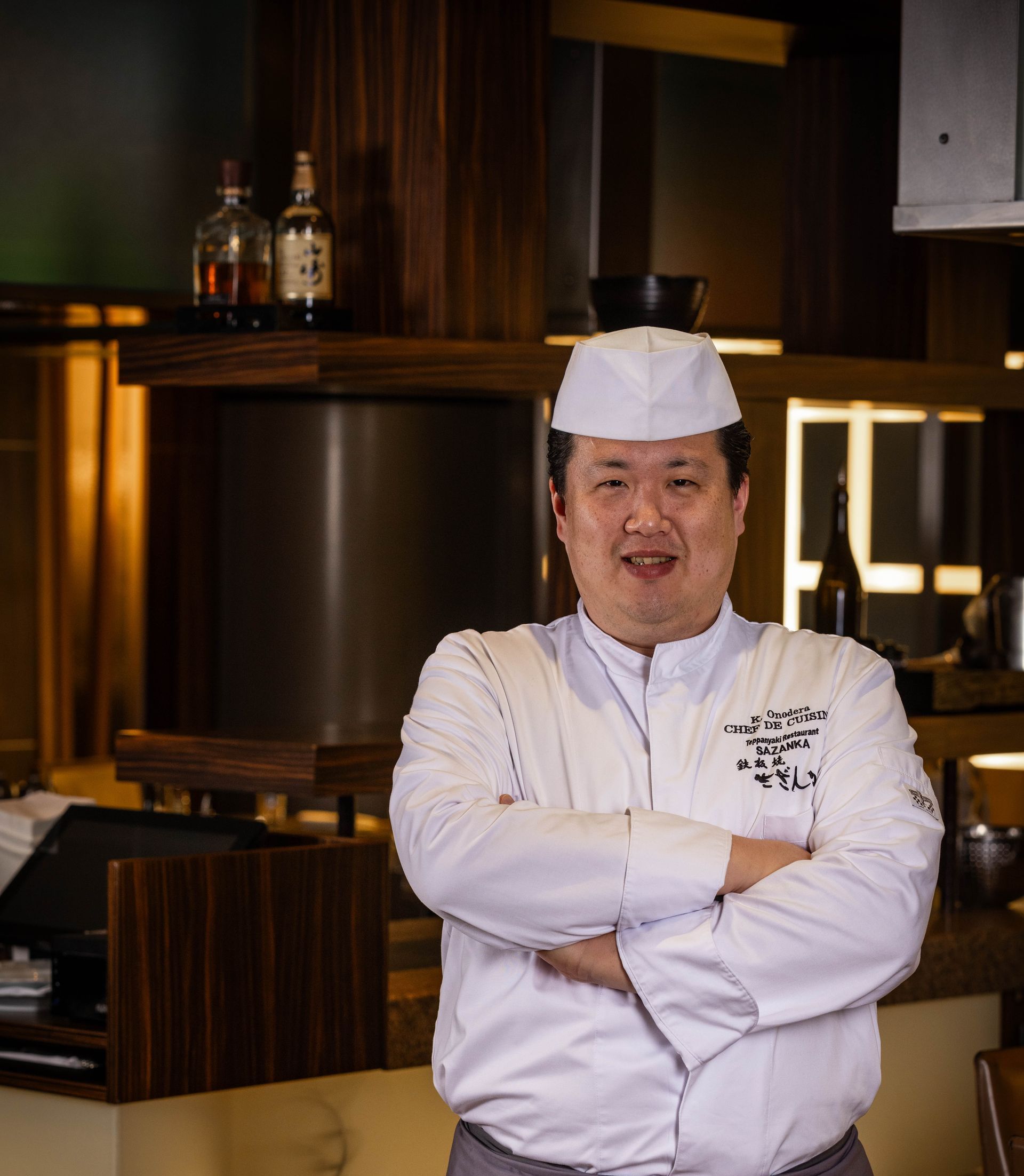 Chef in white uniform, arms crossed, smiles. Inside kitchen setting, dark wood and bottles visible.