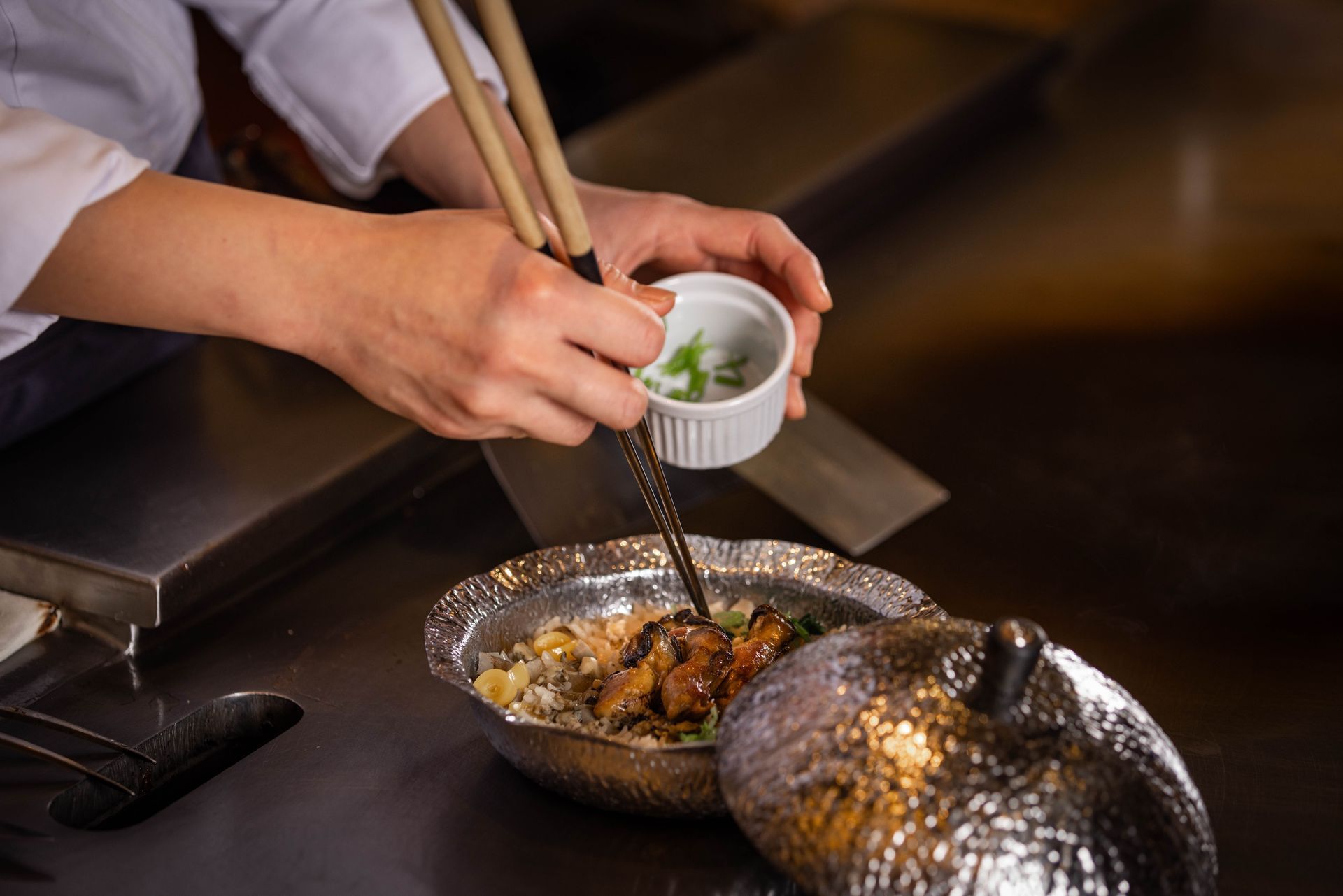 Chef garnishes a stir-fry dish with chopsticks over a teppanyaki grill.