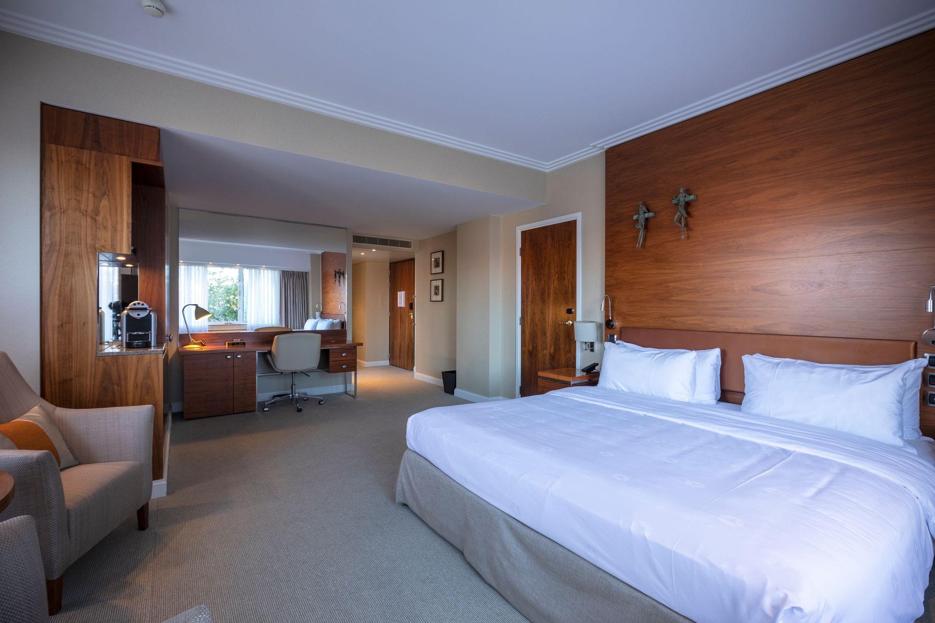 Hotel room with a bed, desk, and chair. Large wooden headboard, window with curtains, and neutral colors.