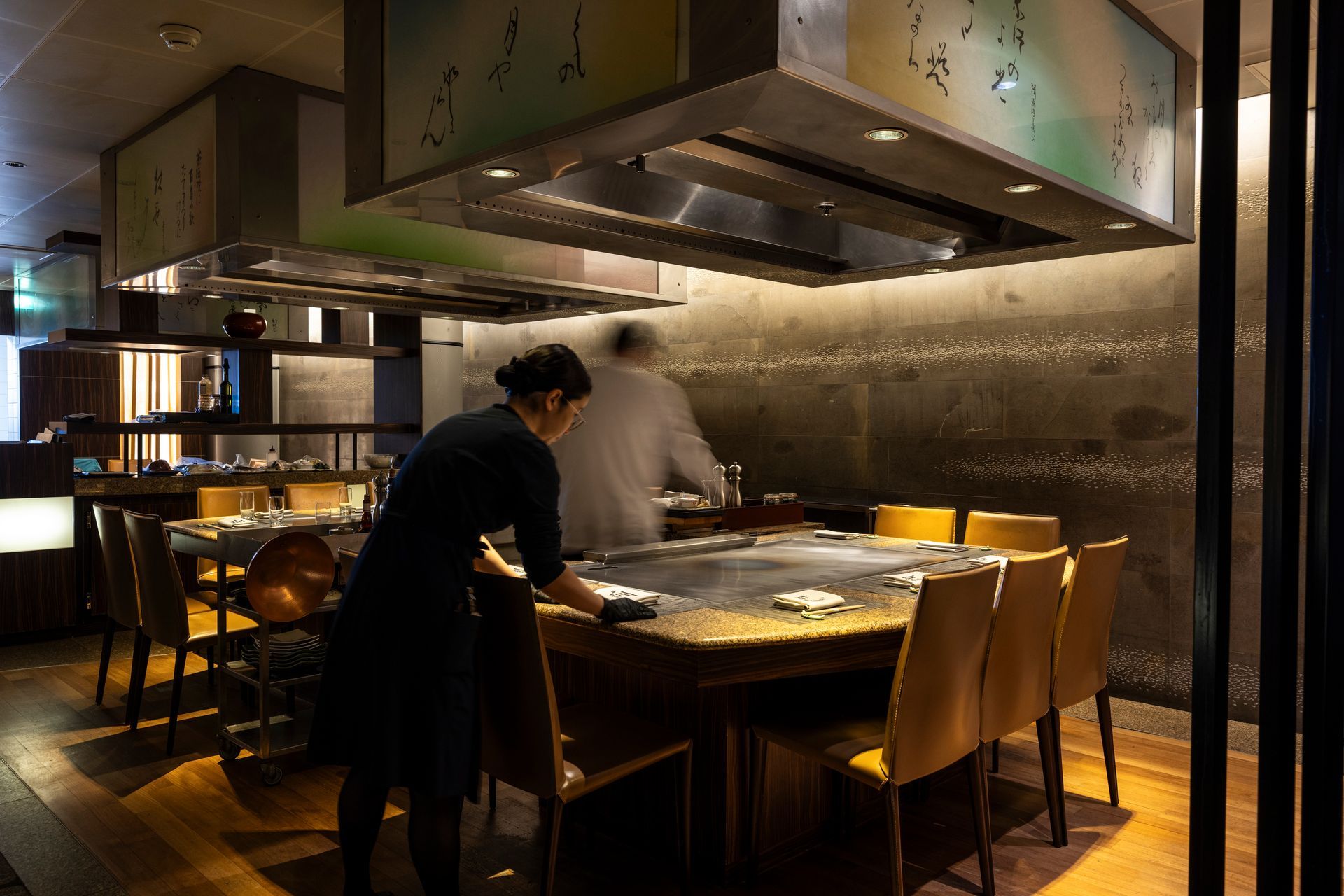 Teppanyaki restaurant: server sets table, chef prepares food at the grill.