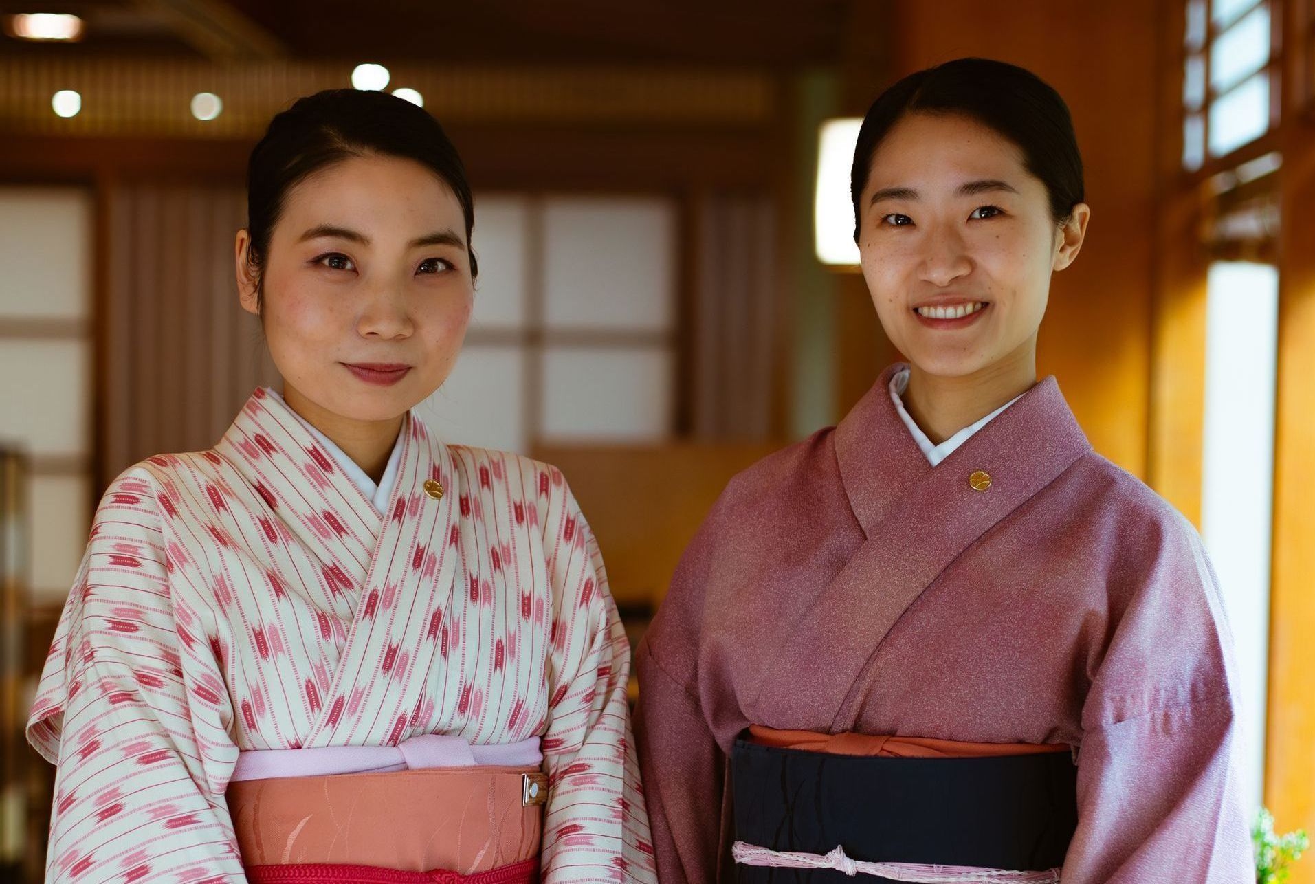 Two women in kimonos pose indoors, smiling. One wears a pink floral pattern, the other a purple kimono.