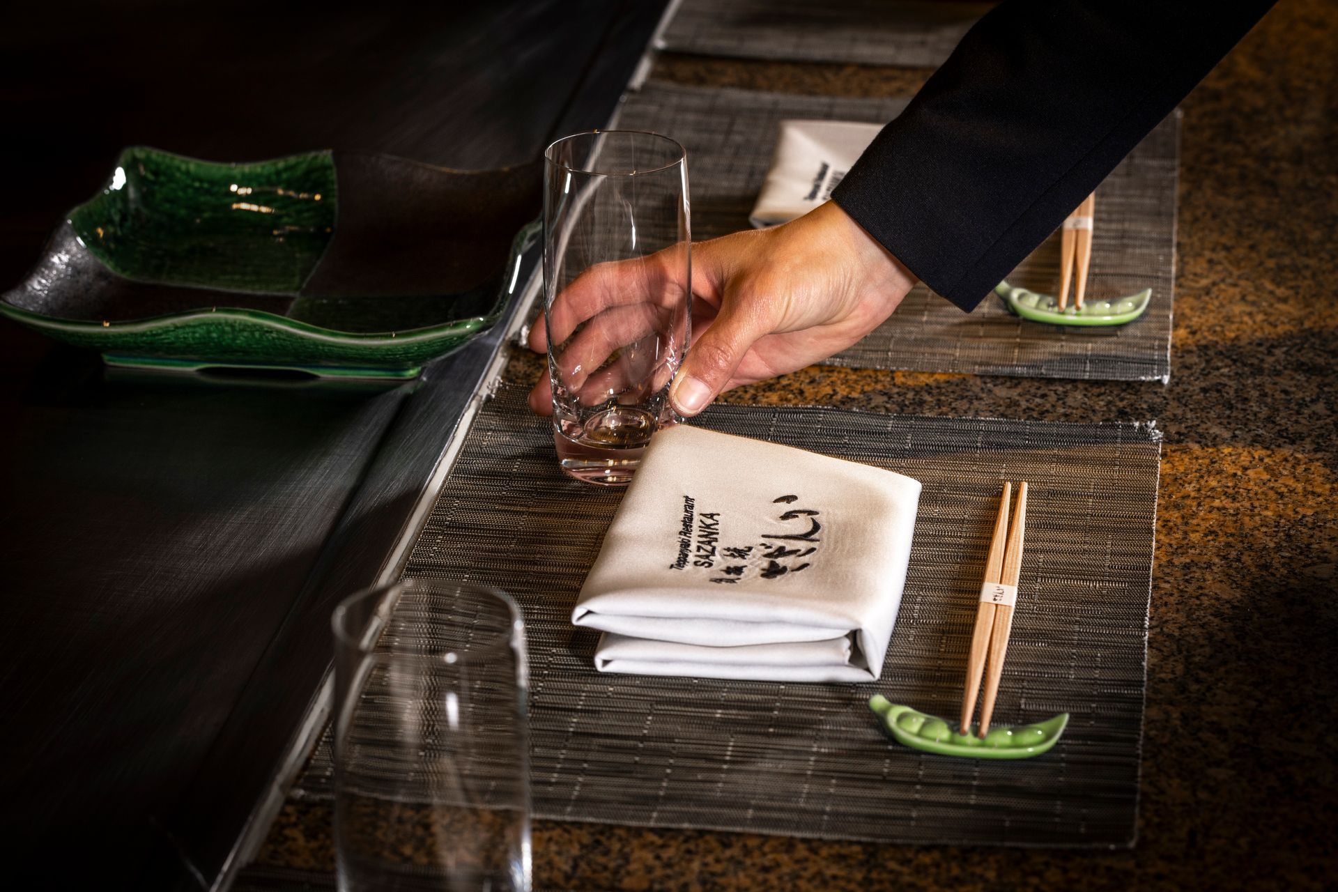 A hand reaching for a glass on a table set for Japanese dining. Chopsticks, napkins, and dishes are present.