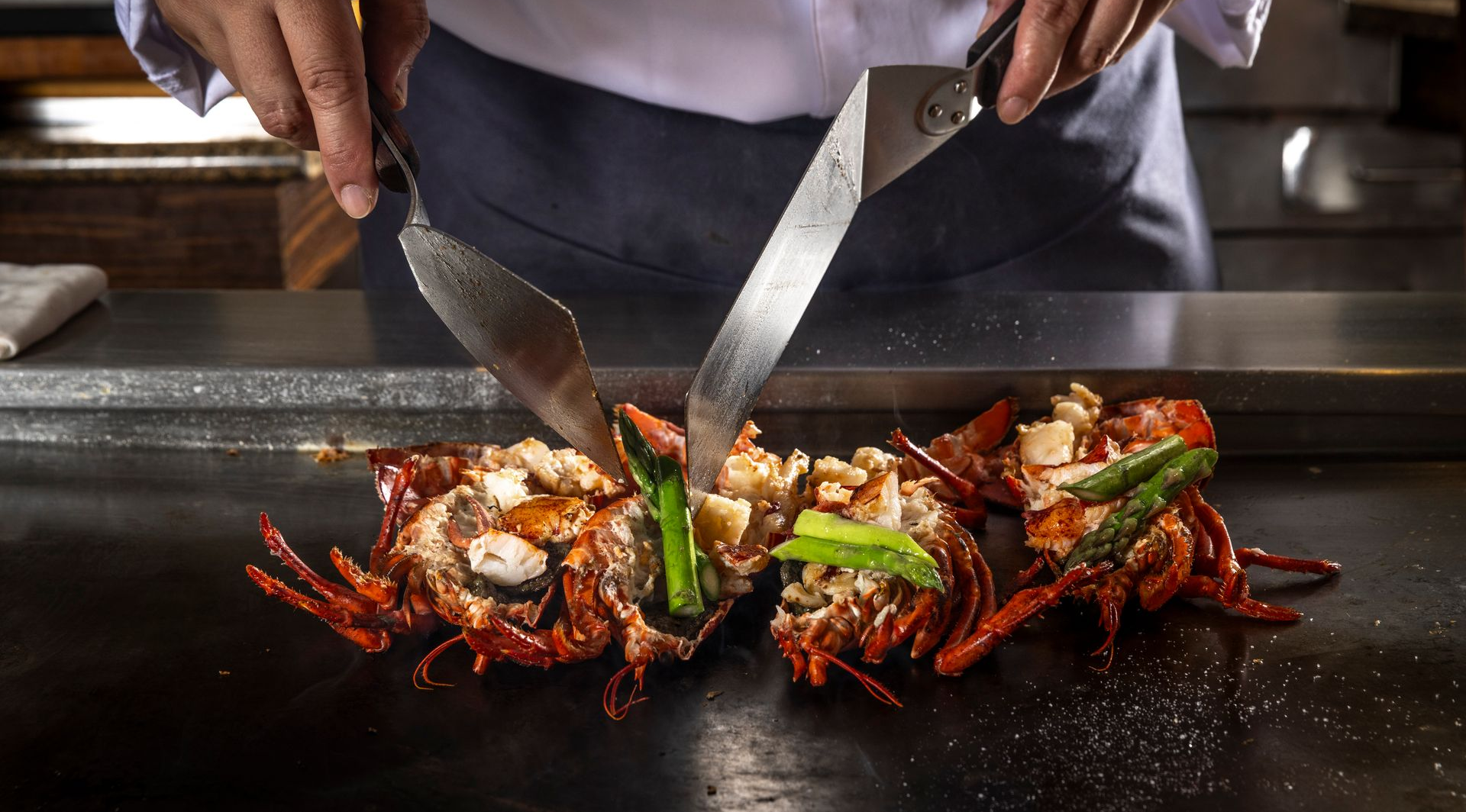 Chef grilling lobster with asparagus on a flat top grill.
