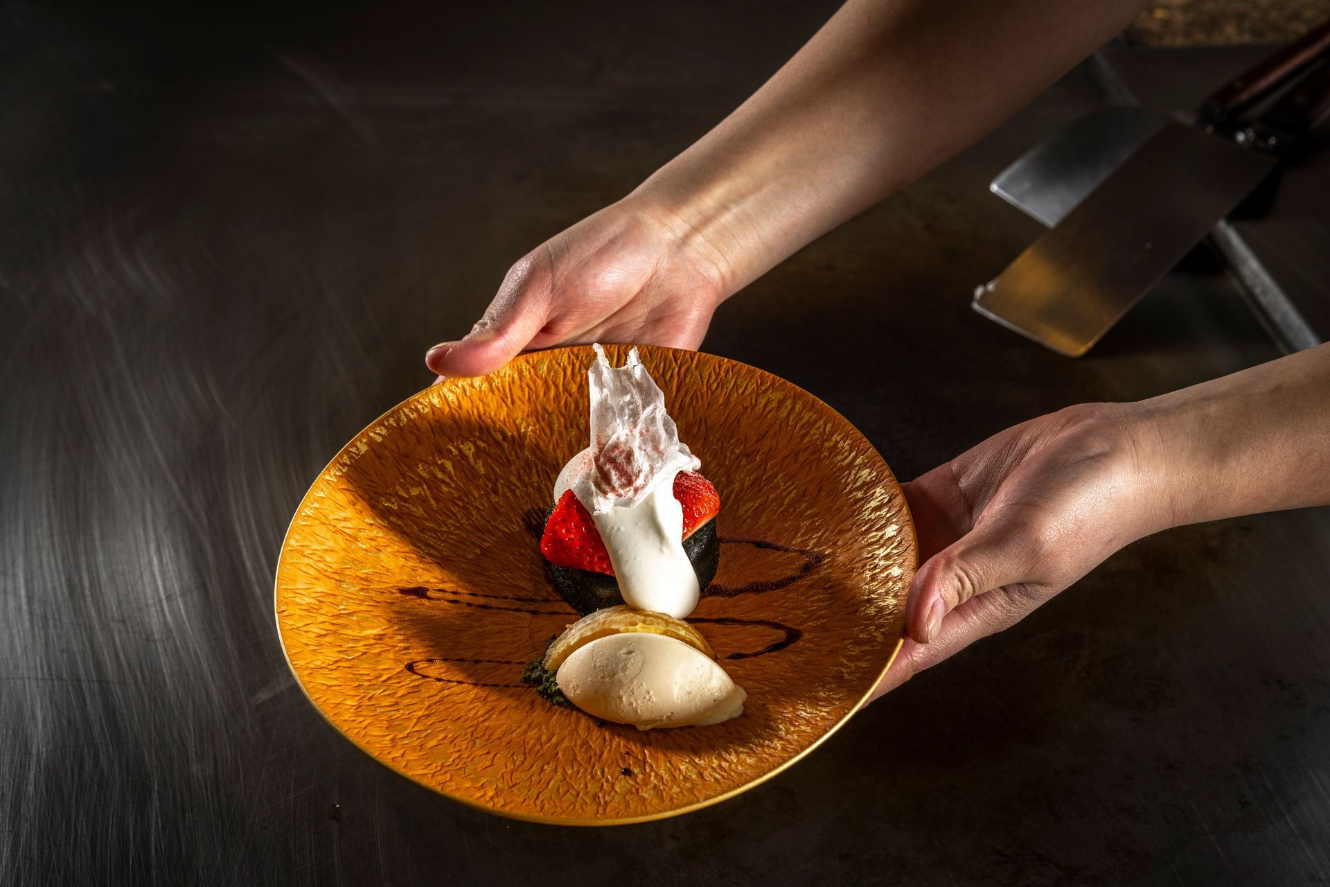 Hands holding a golden plate with a dessert: dark cake, whipped cream, berries, ice cream.
