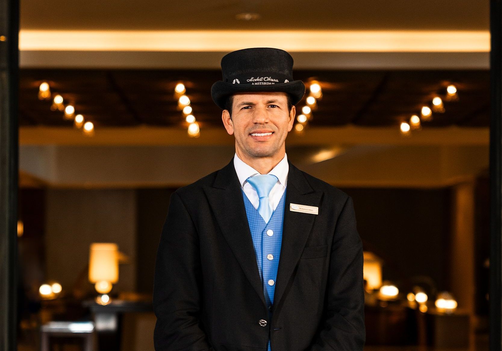 Hotel concierge in uniform, smiling in an entryway. Wearing a hat, vest, and jacket. Warm lighting.