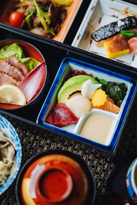 Bento box with various Japanese dishes, including sushi, seafood, and salad, presented on a dark surface.