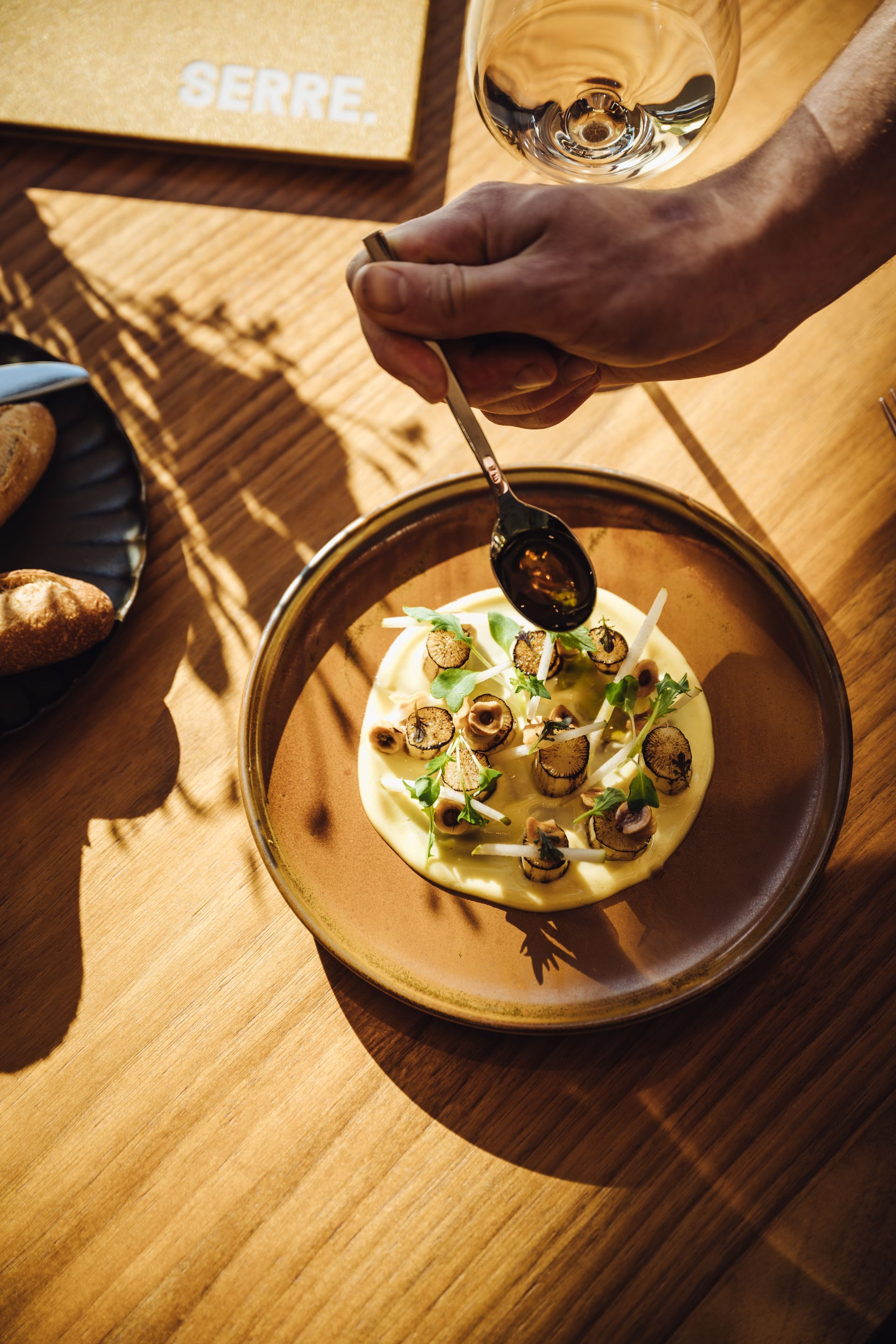 Person using knife and fork to eat a plate of food, with a glass of wine on a wooden table.