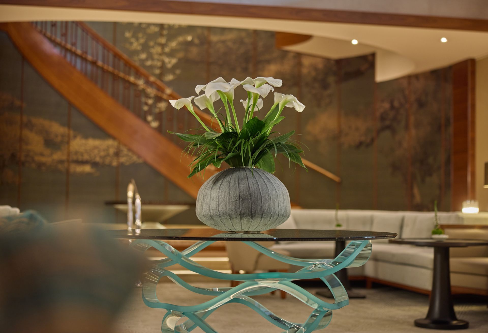 White calla lilies in a textured vase on a glass-topped table. A grand staircase and lounge area are in the background.