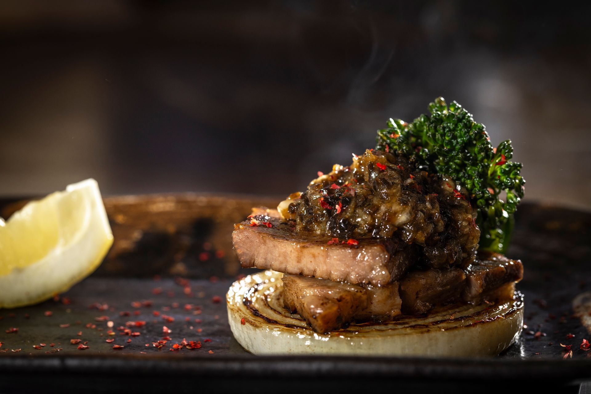 Steak with sauce and broccoli on a grilled onion, served on a dark plate, with a lemon wedge.