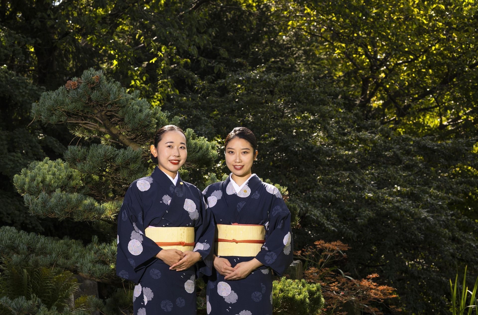 Two women in yukata stand in front of a green, leafy background.