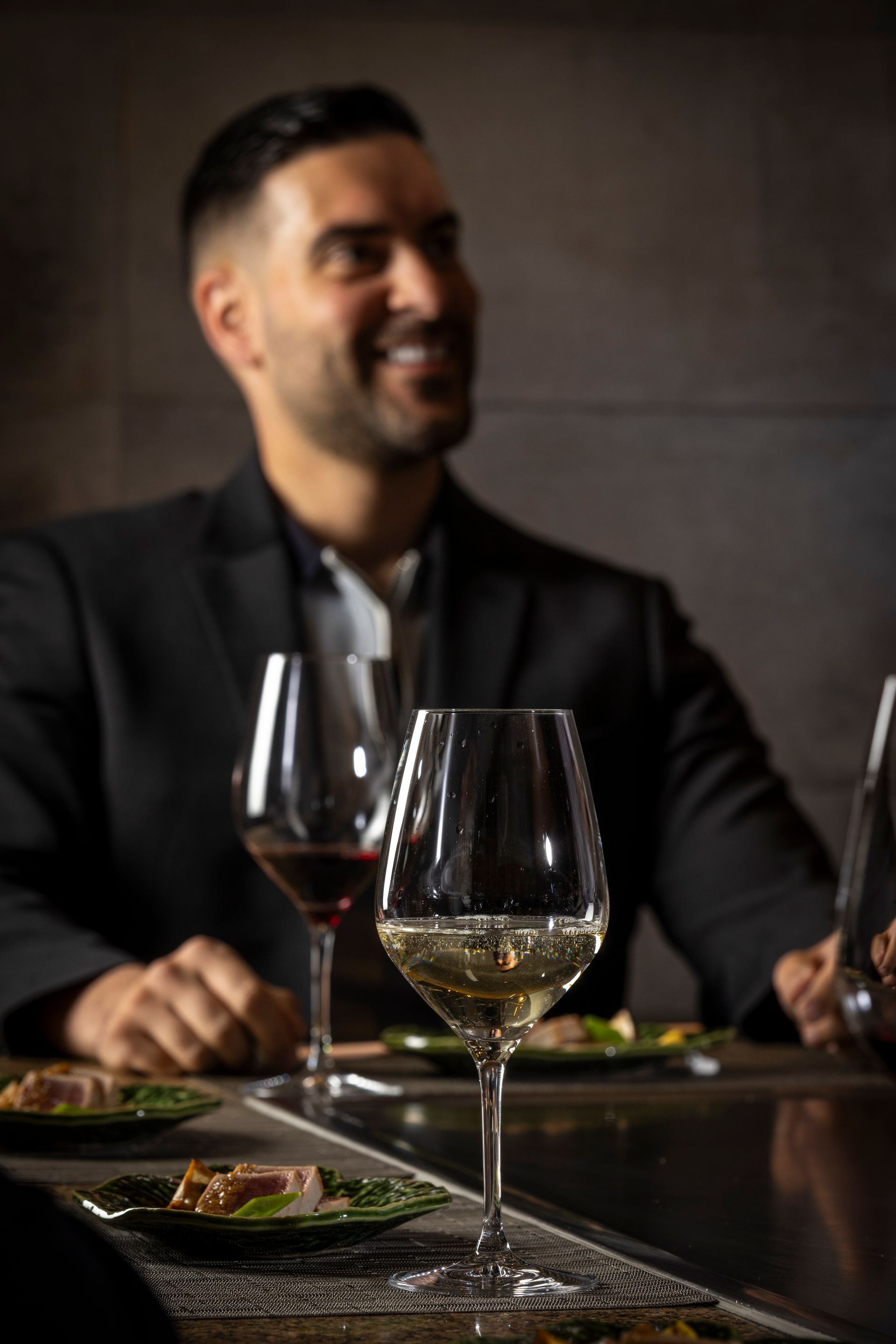 Man smiling at a table with wine glasses, featuring red and white wine. Plates of food are also on the table.