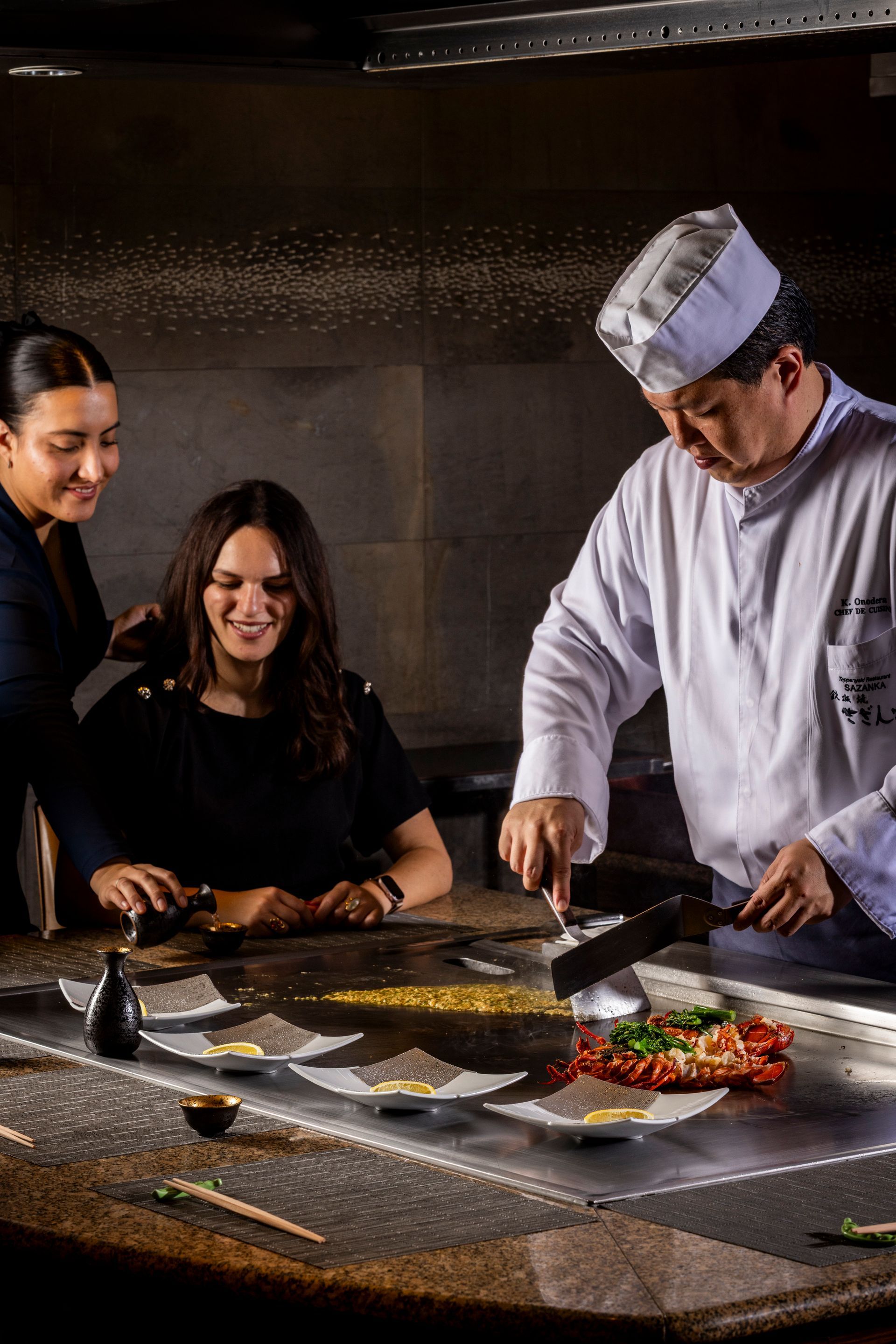 Chef cooking at a hibachi grill, serving two women. Sparks fly, seafood and plates are visible.