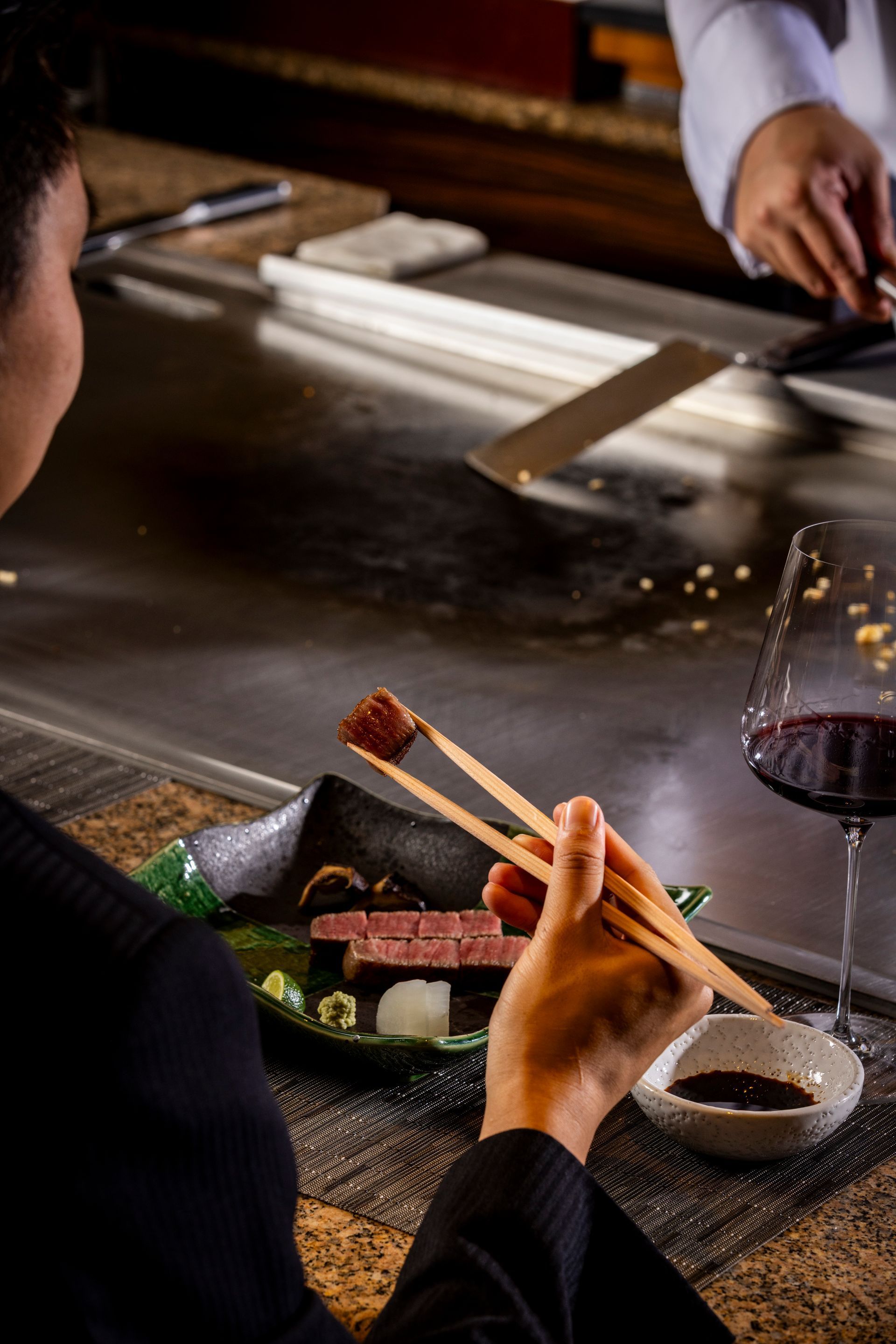 Person eating steak with chopsticks, teppanyaki chef in background, red wine, elegant setting.