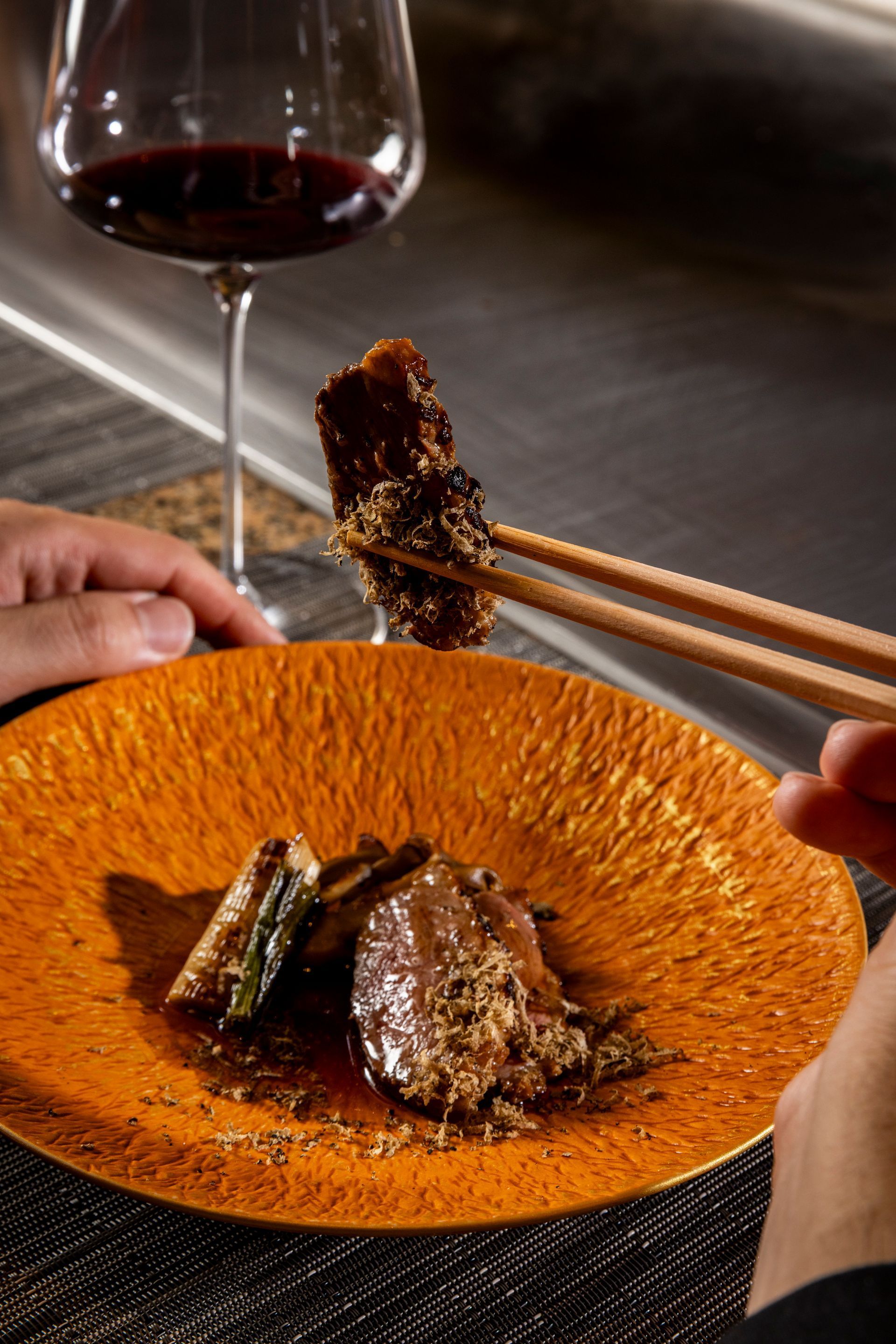 Person using chopsticks to pick up food from orange plate; wine glass in the background.