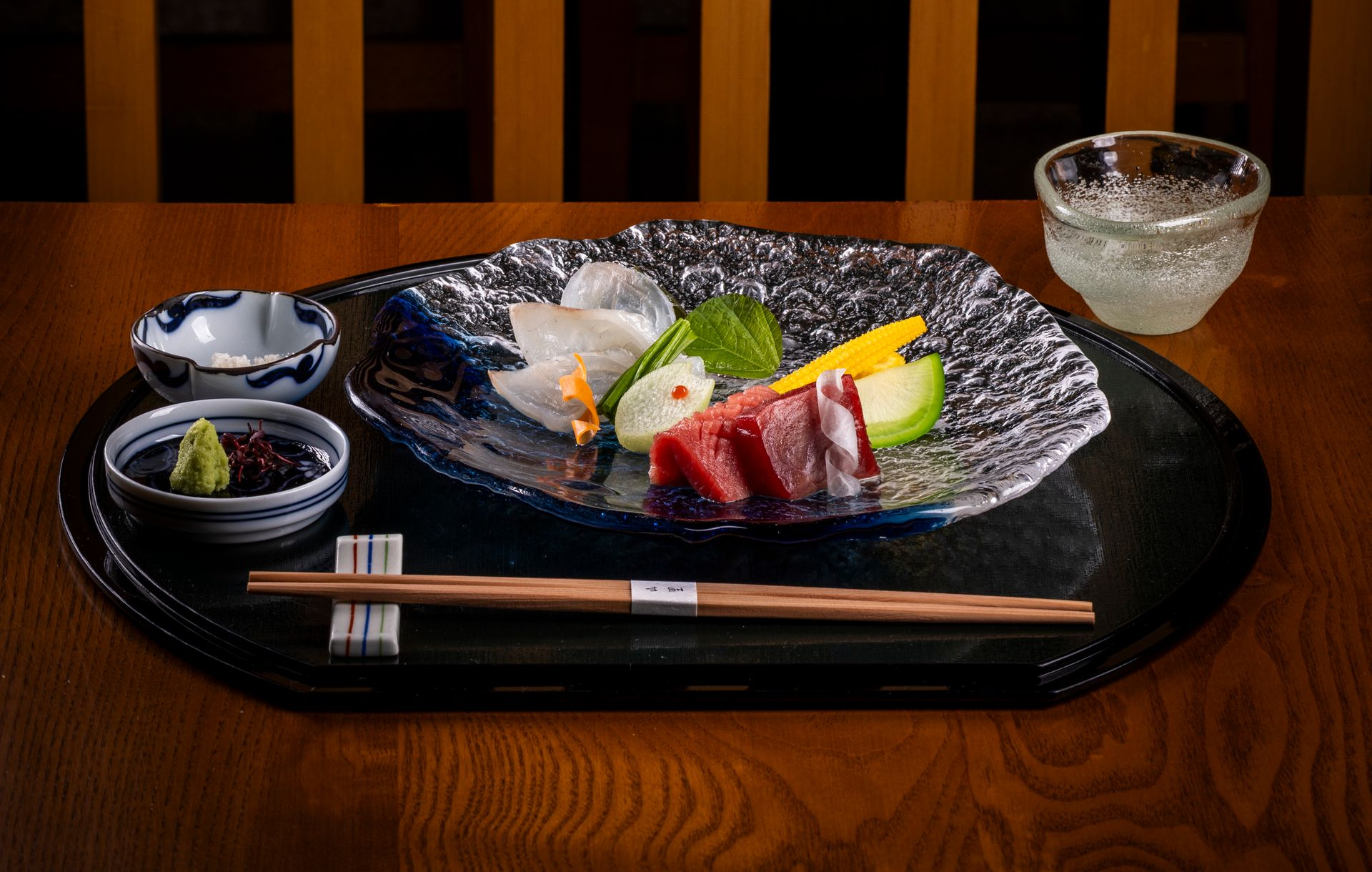 Sushi plate with sashimi, soy sauce, wasabi, and chopsticks on a wooden table.
