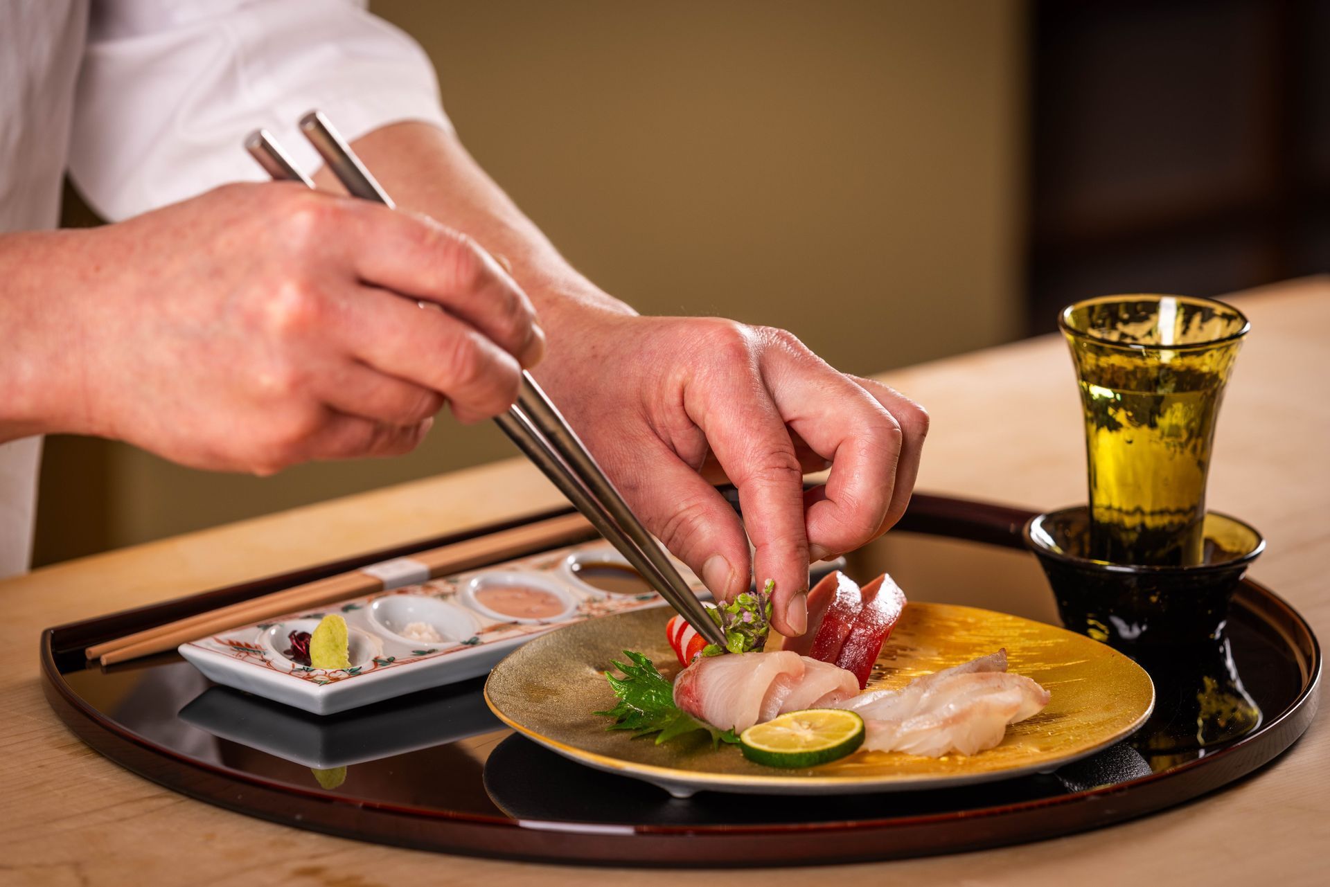 Chef arranging sashimi on a golden plate with chopsticks, sake glass nearby.