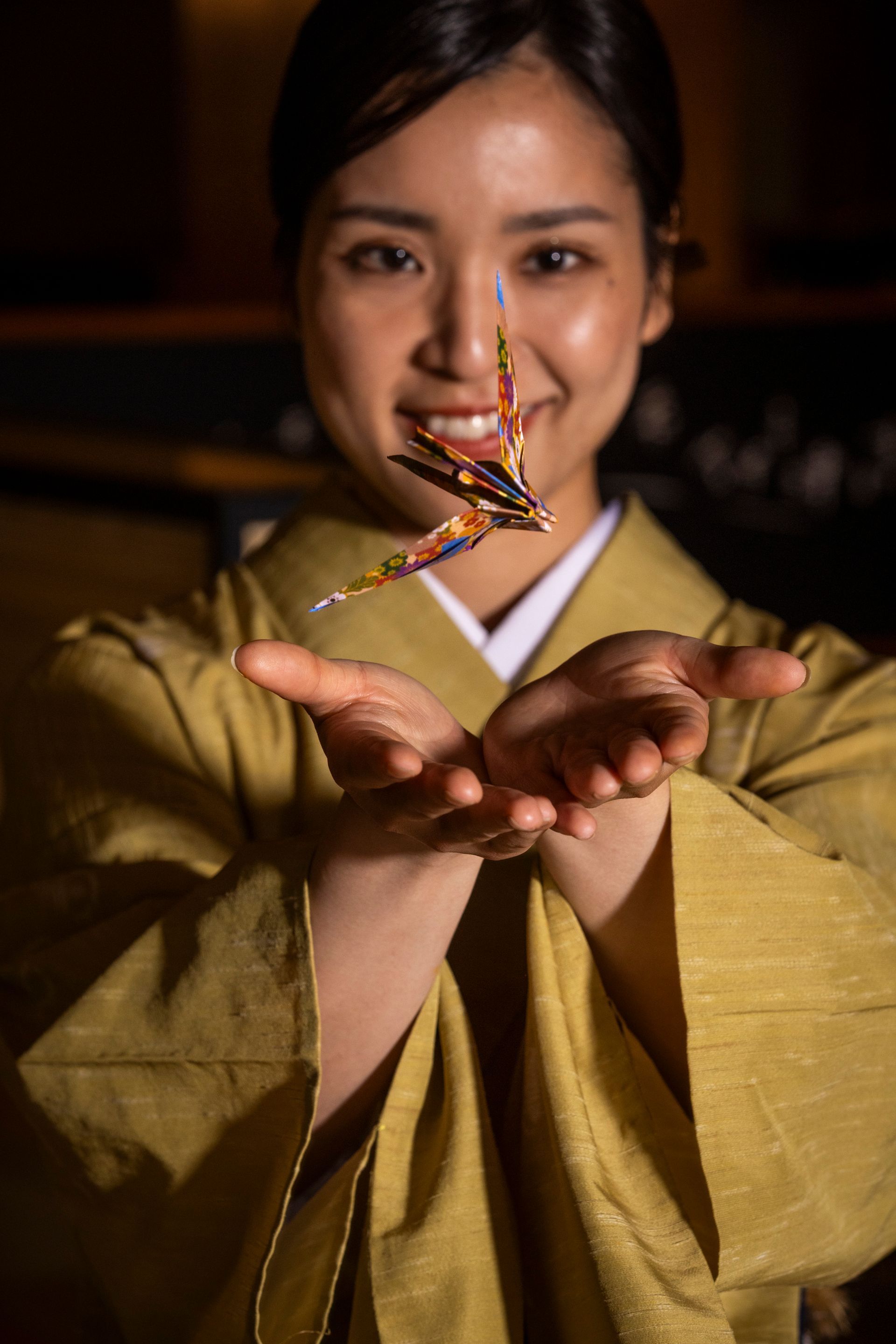 Woman in gold kimono smiles, holding a colorful origami crane in her hands.