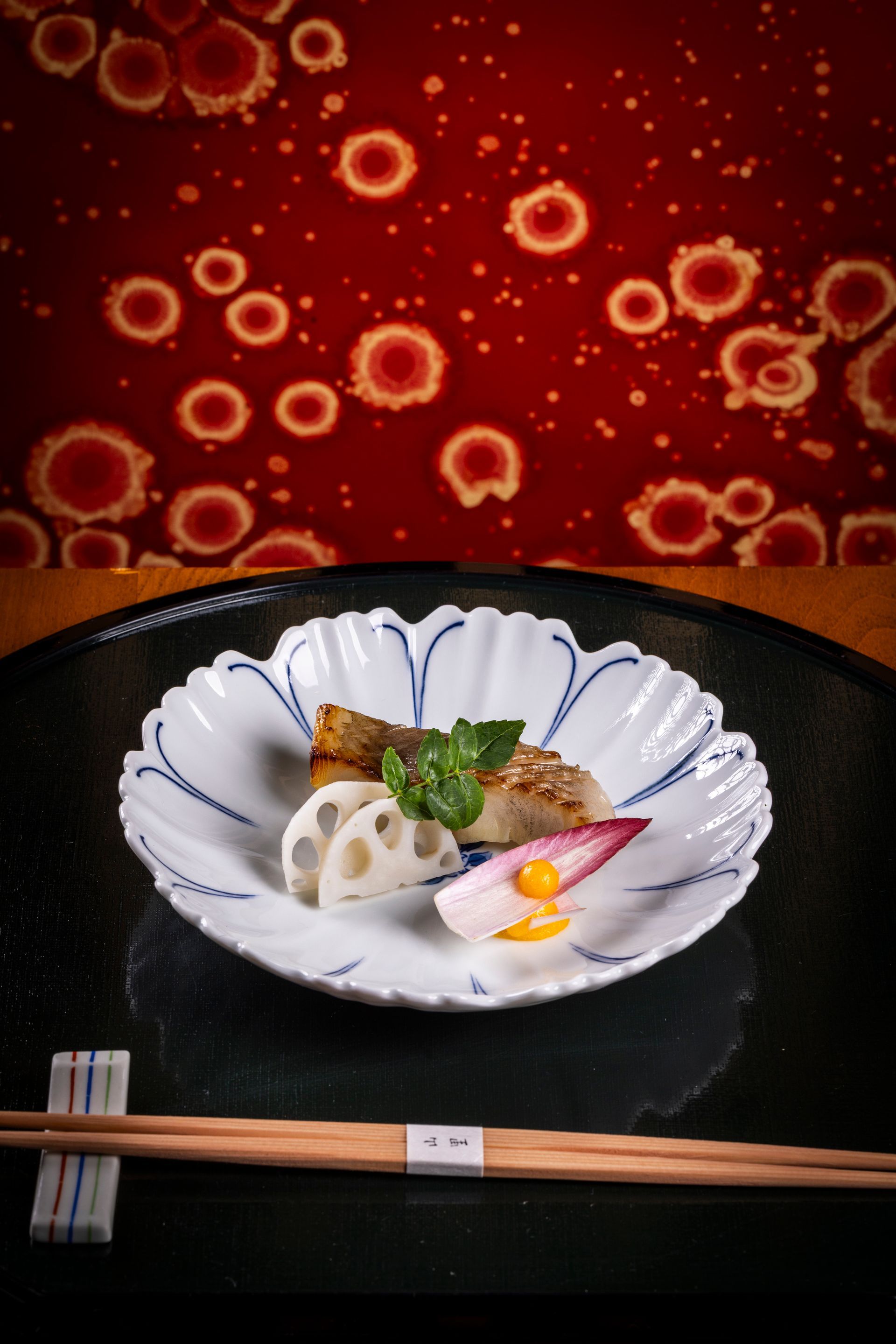A Japanese dish with fish and vegetables on a white, petal-shaped plate, chopsticks. Red background with white circles.