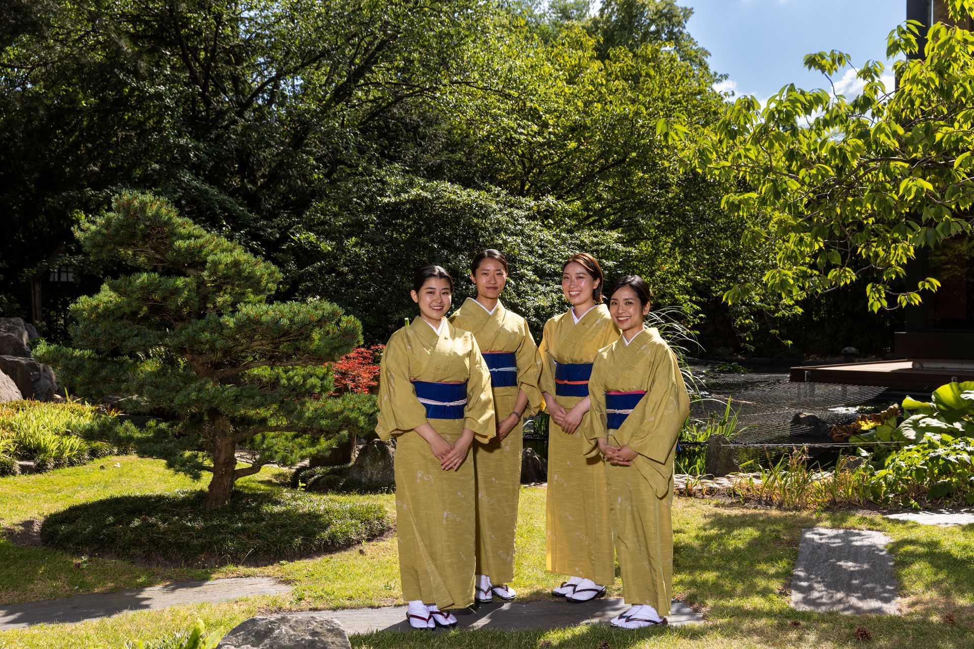 Four people in yellow kimonos stand in a Japanese garden with trees and foliage.