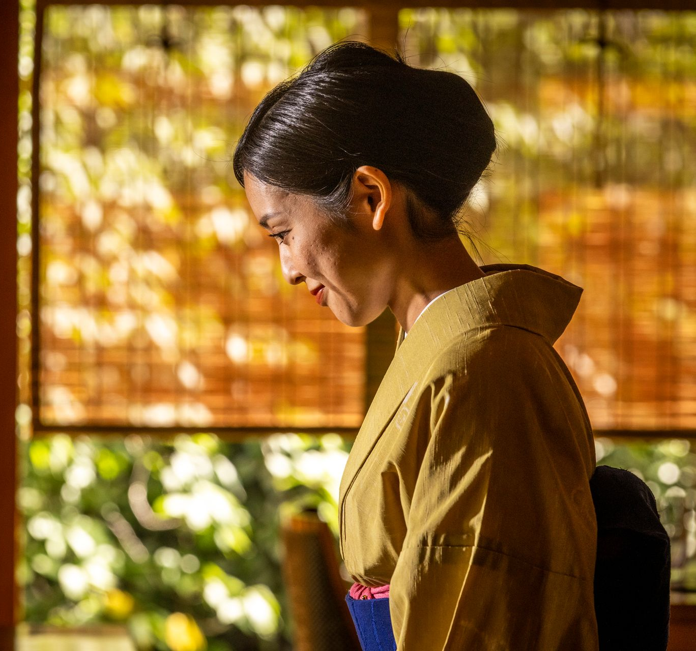 Woman in kimono bows head, lit by sunlight through blinds.