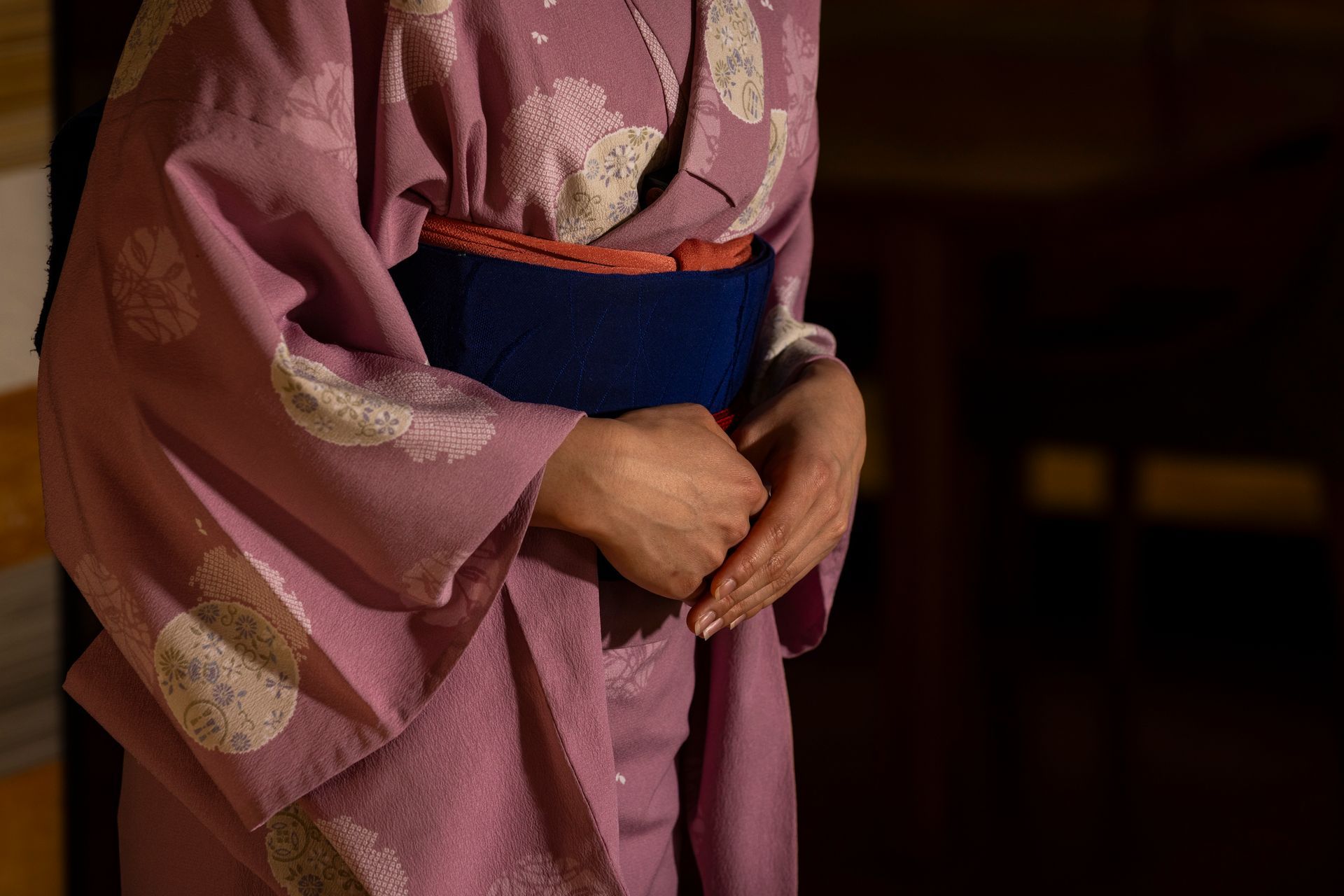 Woman in pink kimono with blue obi, hands clasped over stomach.