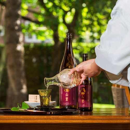 Person pouring sake from a carafe into a glass, two bottles, and greenery in the background.