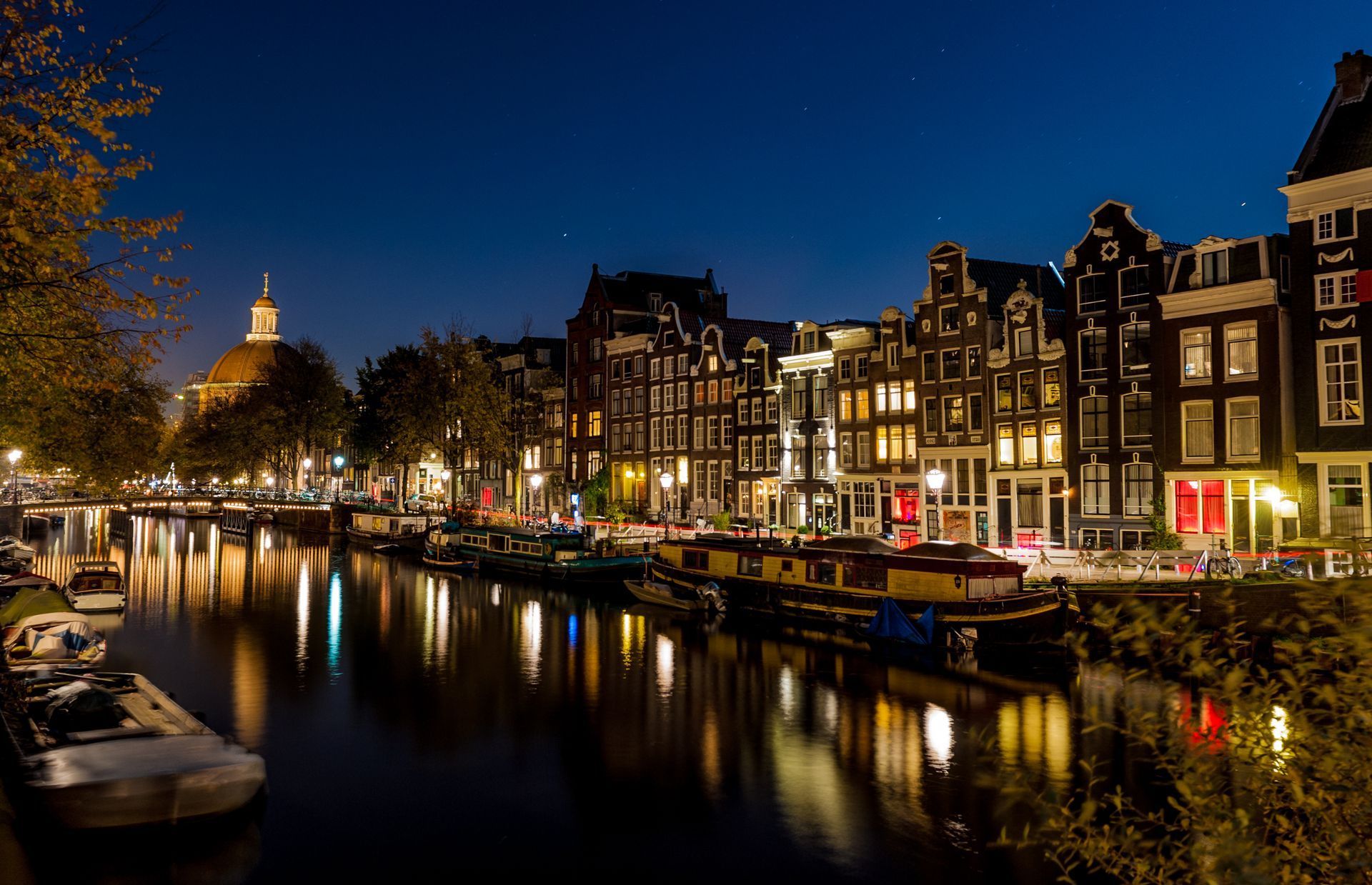 Night view of Amsterdam canal with lit buildings, boats, and reflecting water under a dark blue sky.