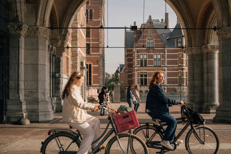 Two people on bikes ride under an arched walkway with a building in the background.