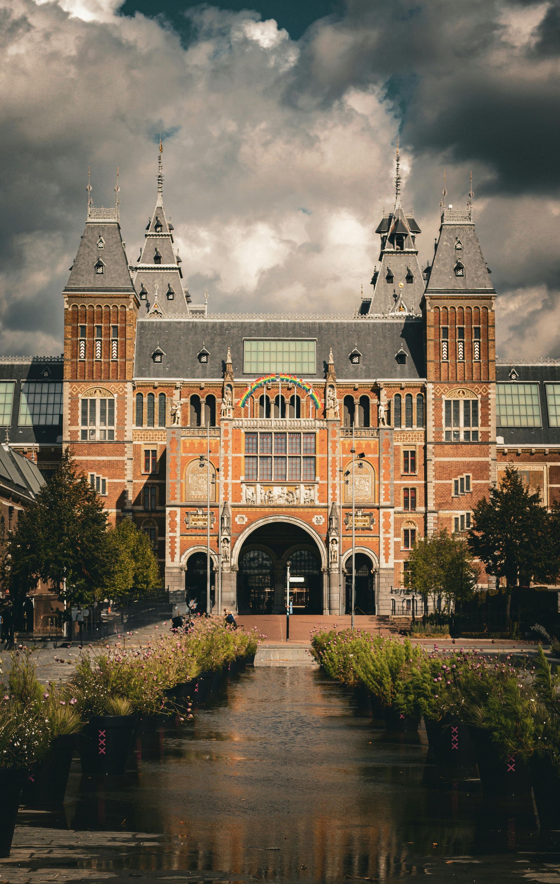 Rijksmuseum, Amsterdam: sierlijk bakstenen gebouw met torens en een boogvormige ingang, een bewolkte lucht erboven, weerspiegeld in een gracht.