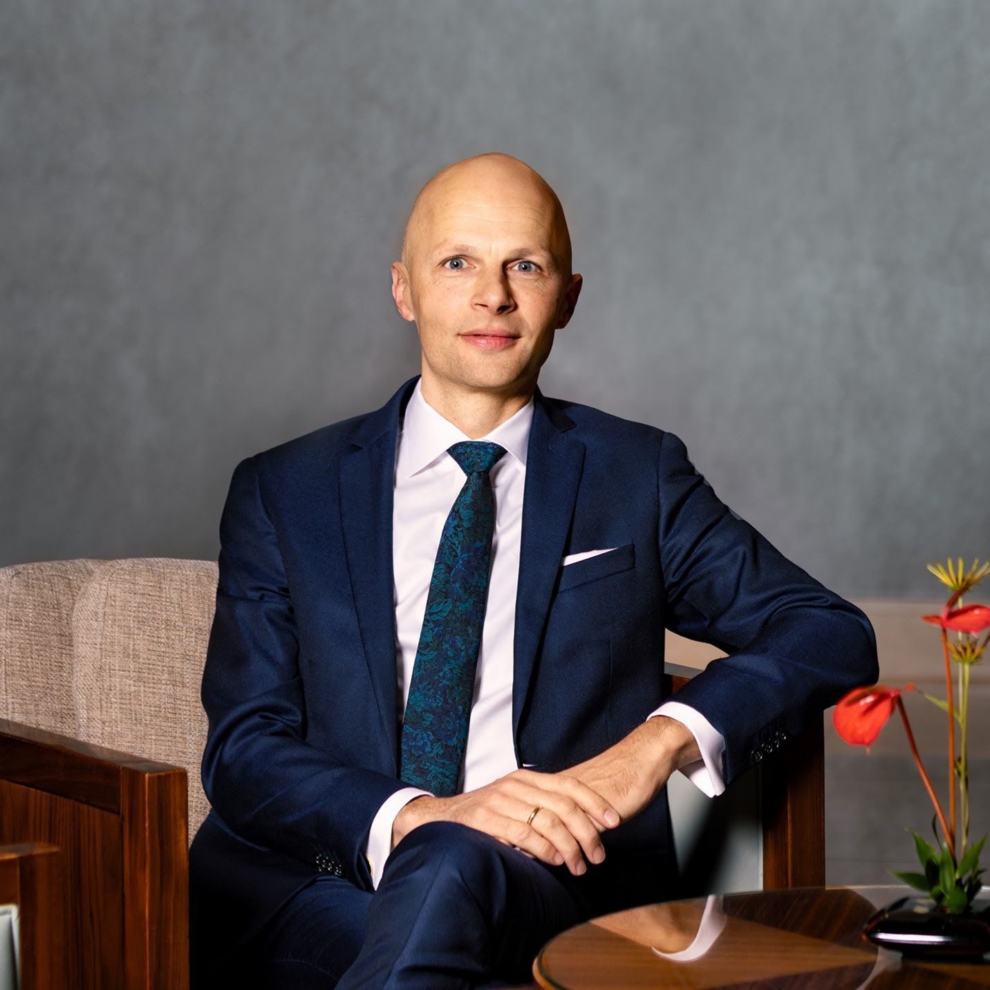 Bald man in a blue suit, sitting, looking forward, small red flowers on right.