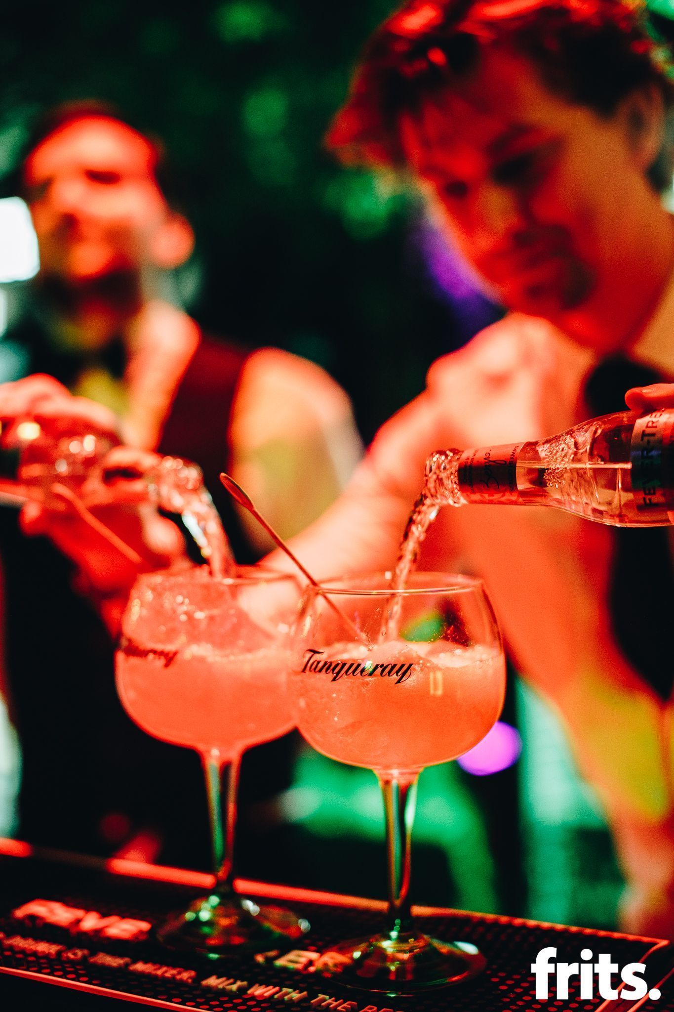 Two bartenders pouring drinks in a dimly lit bar; cocktails in the foreground.