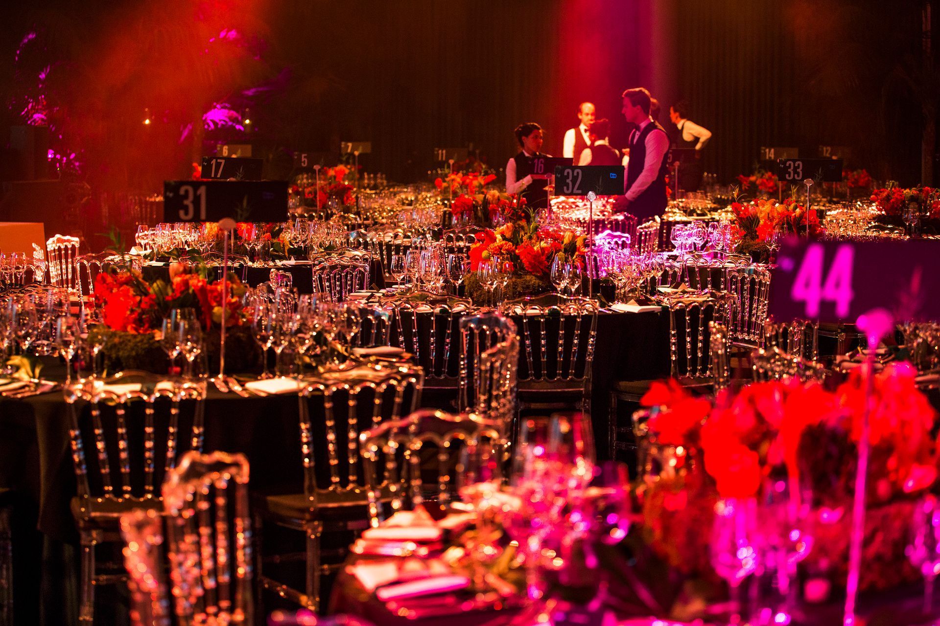 Tables set for a gala dinner, with clear chairs, dark linens, and red floral centerpieces. Servers in background.