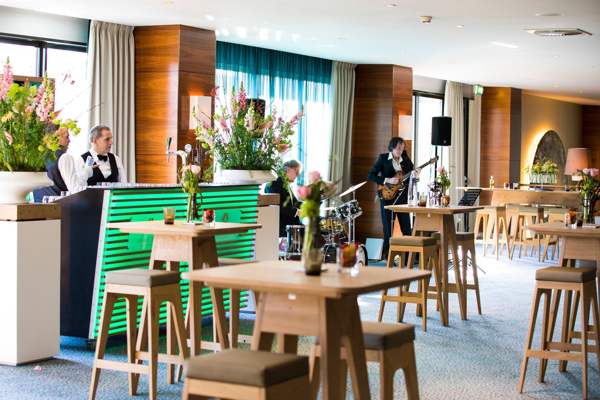 A band performs in a modern lounge. Servers behind bar, tables, and flowers.
