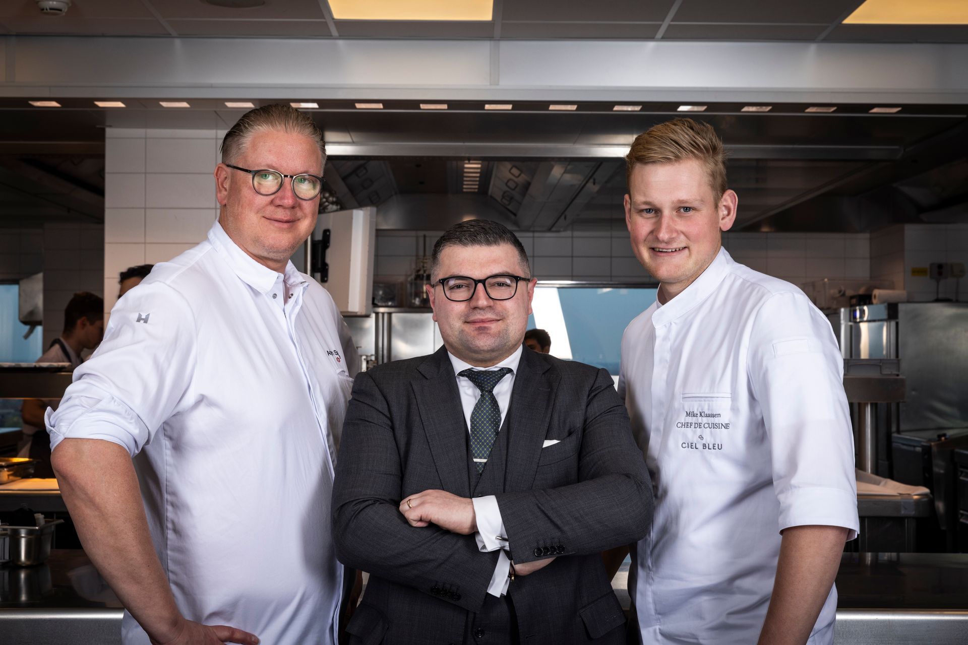 Three people in a modern kitchen. A man in a suit stands between two men in white chef's coats.
