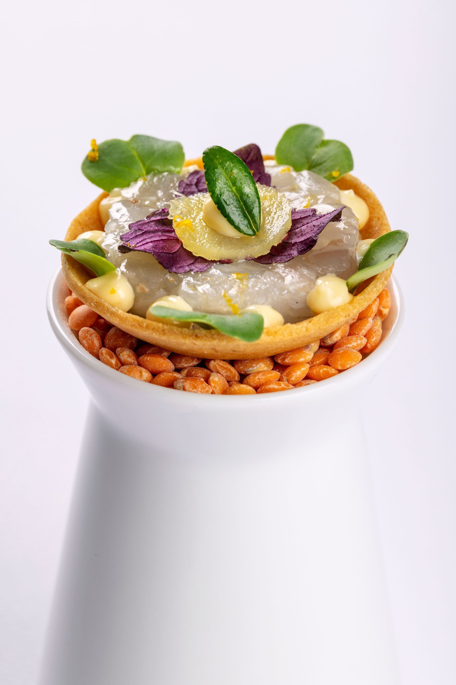 Gourmet dish in a white stand. Contains shrimp, purple cabbage, green leaves, and orange rice crackers.