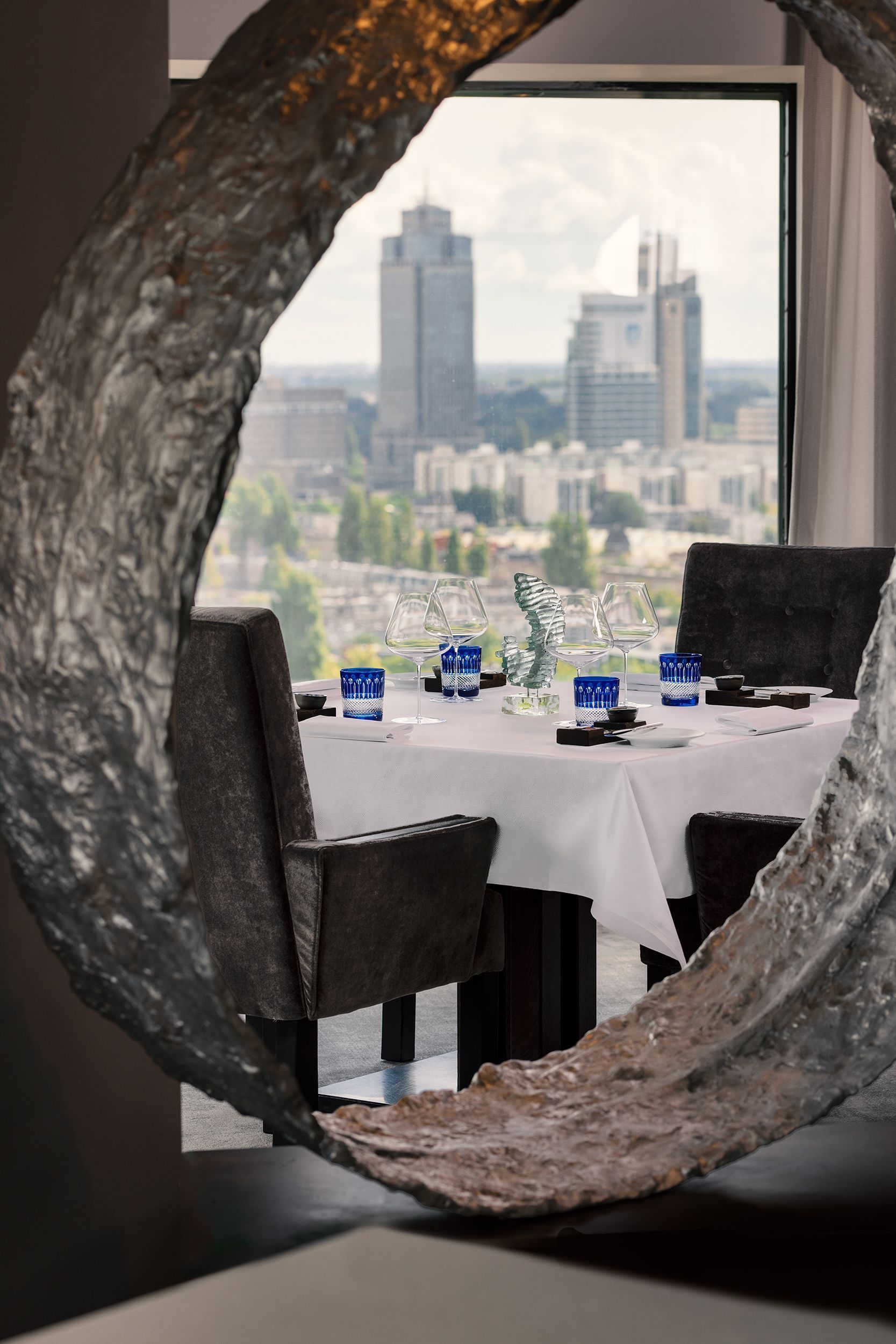 Restaurant table set for dining, framed by a textured, circular sculpture; cityscape visible through window.