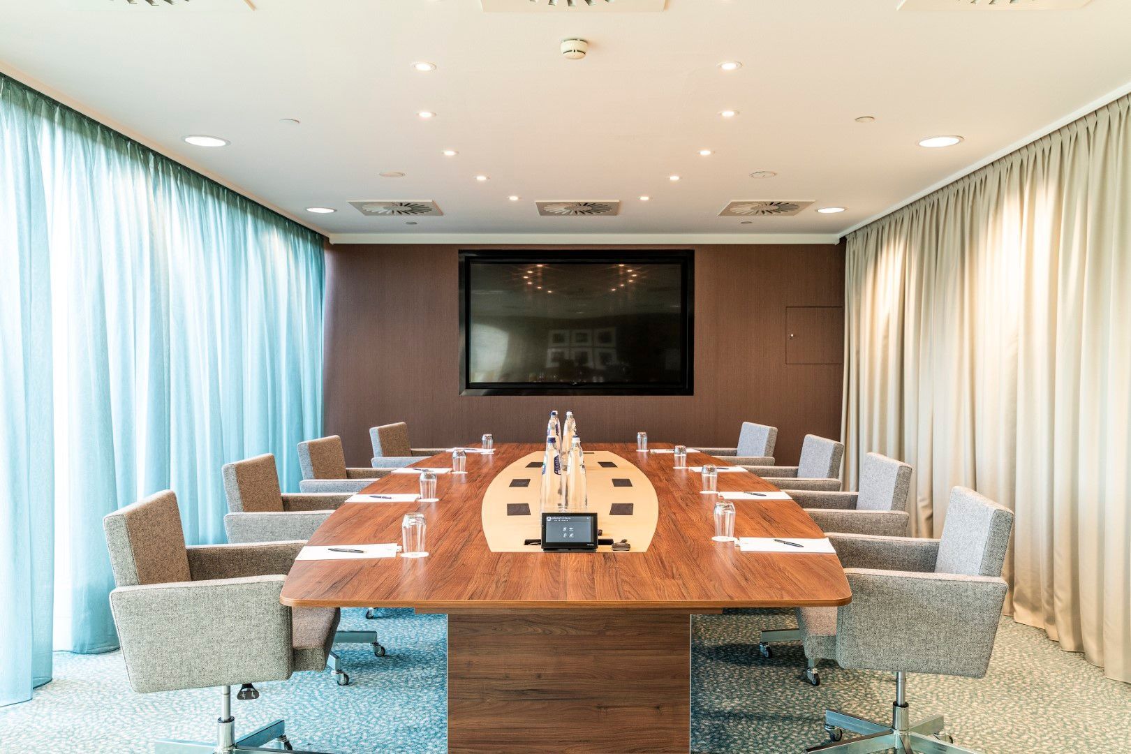 Meeting room with a large wooden table, chairs, and a large screen. Light blue curtains and beige walls.