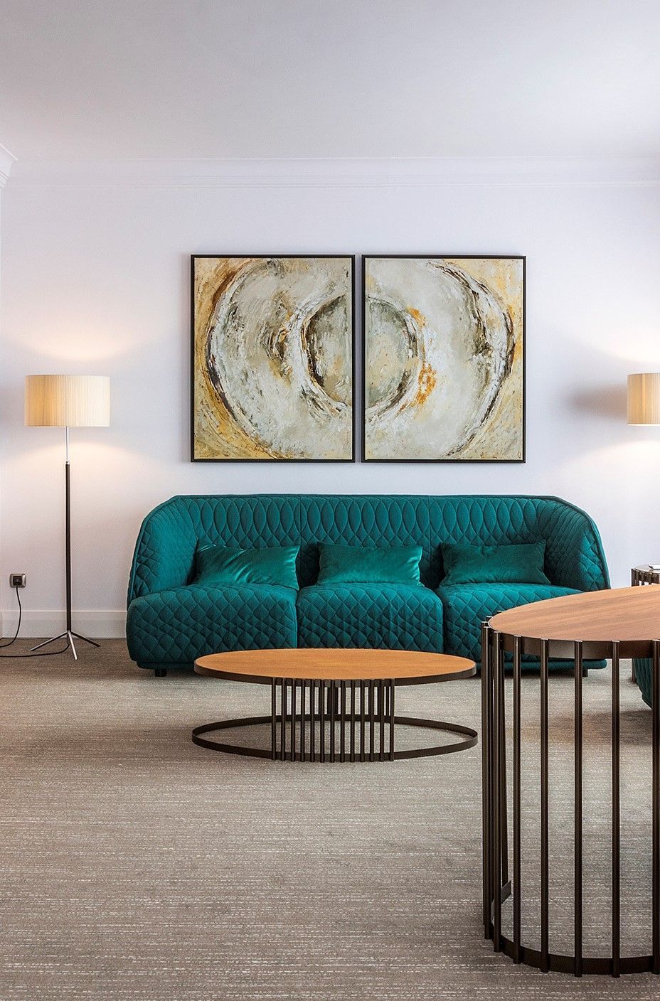 Teal sofa in front of two abstract paintings; a coffee table and side table in front of a neutral-colored carpet.