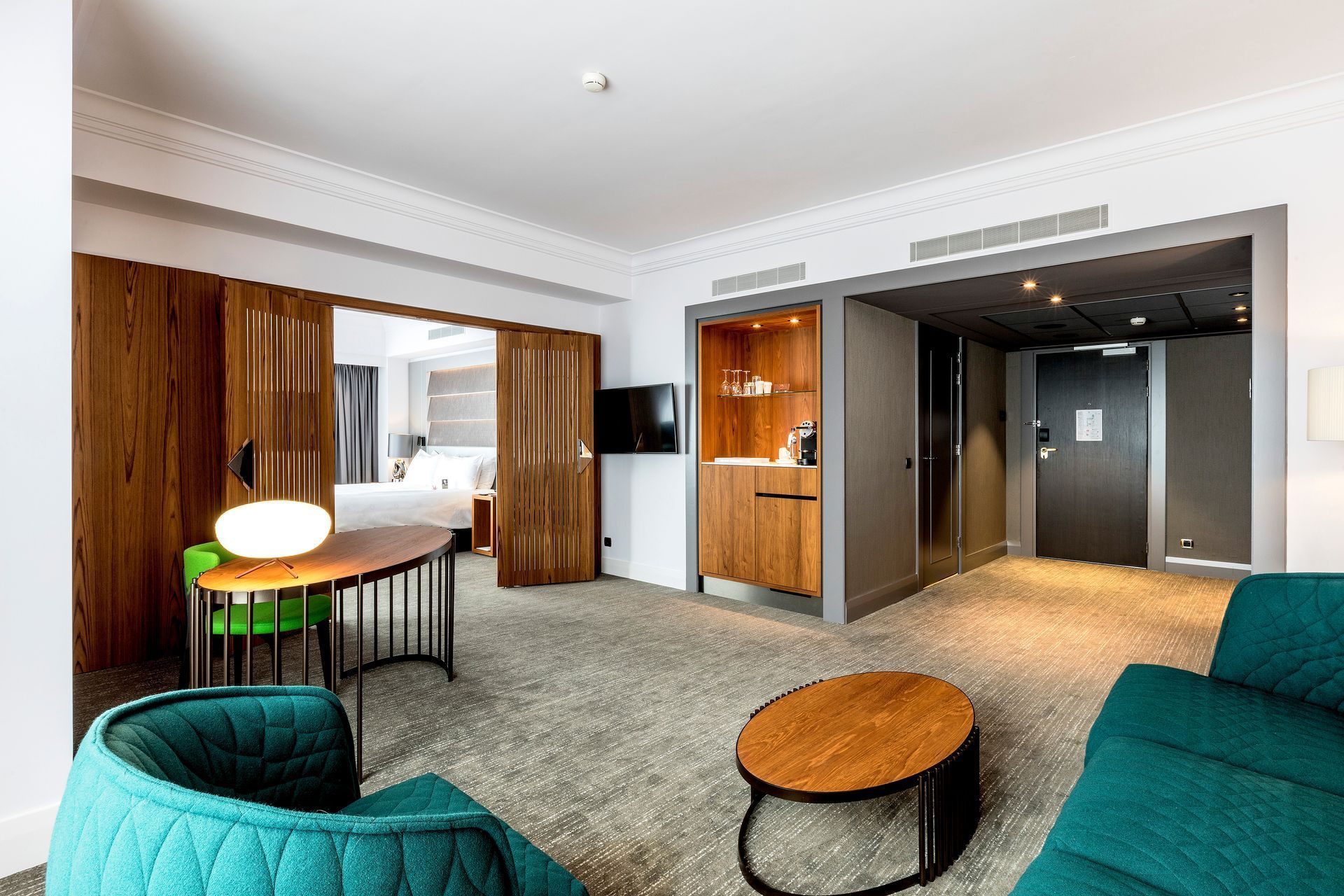 Hotel suite with teal seating, wooden table, and a bedroom visible through open doors.