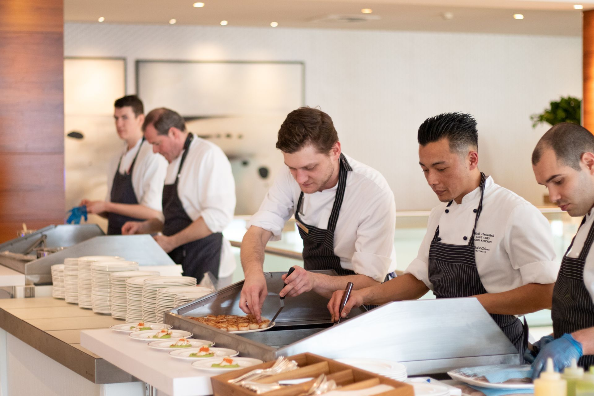 Chefs preparing food at a catering event. Several people in white coats and aprons working on a counter.
