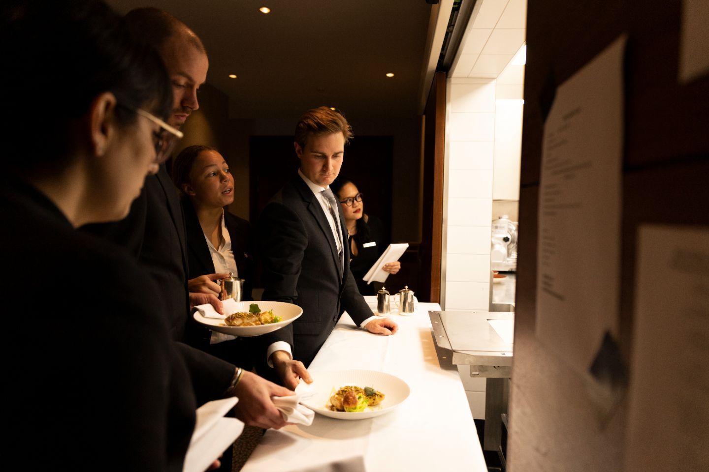 People in suits near a kitchen counter, holding plates of food. Some look toward the food.