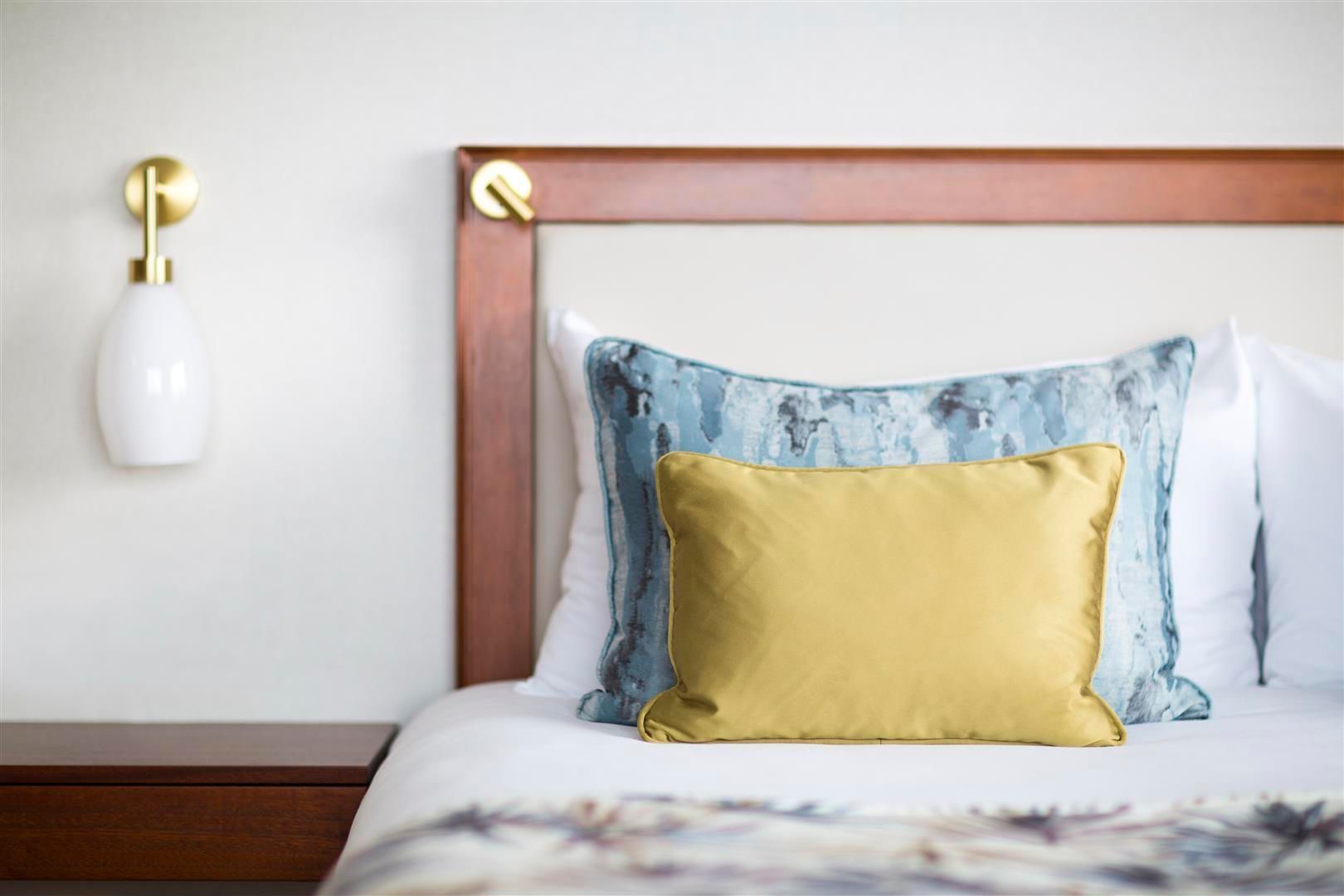 Bed with white bedding, blue and yellow pillows, brown headboard and nightstand, white wall with wall lamp.