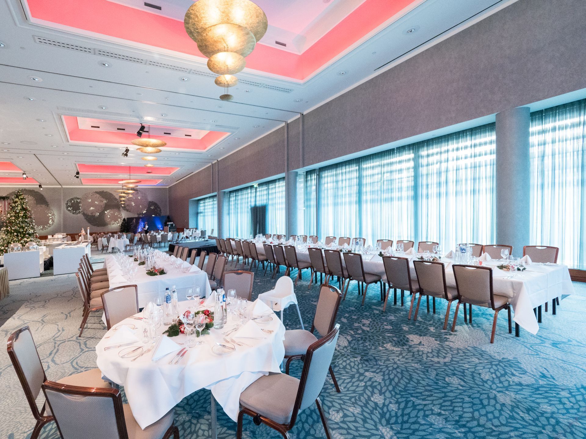 Banquet hall with tables set for a meal, tall windows, and decorative lighting.