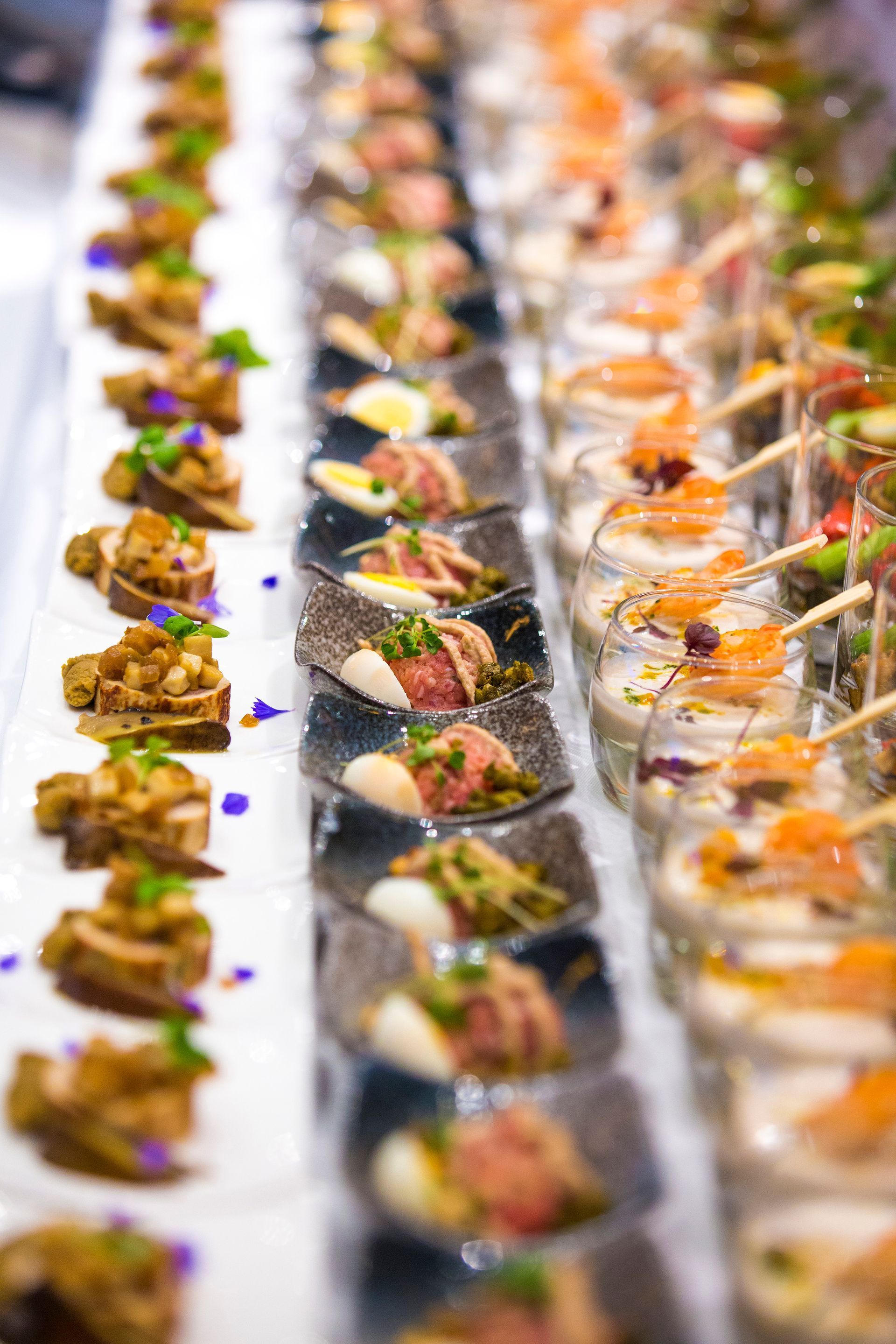 Rows of small, ornate appetizers on a buffet. Various colorful ingredients and textures are visible.
