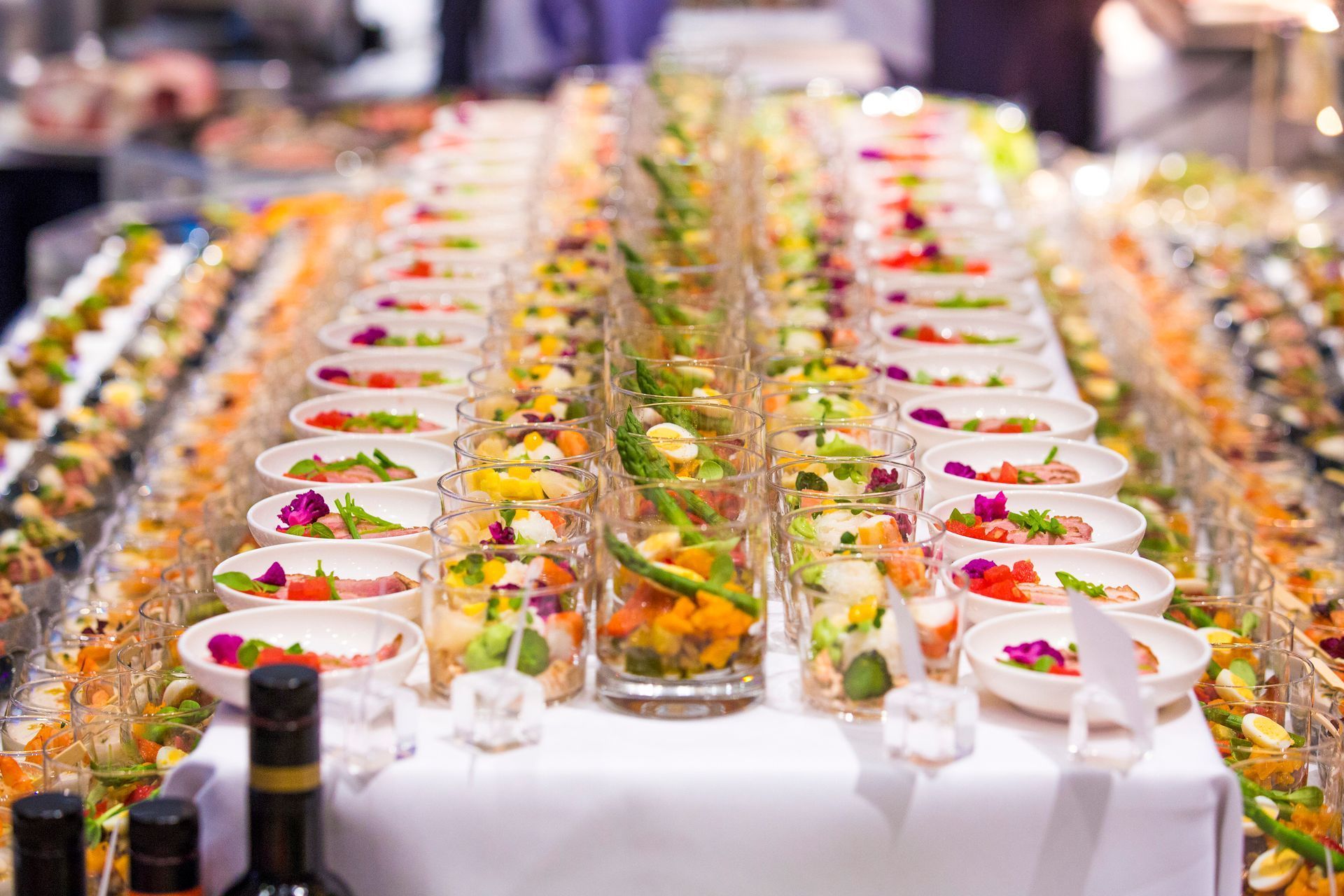 Buffet table with rows of colorful appetizers in clear cups, asparagus garnish, white tablecloth.